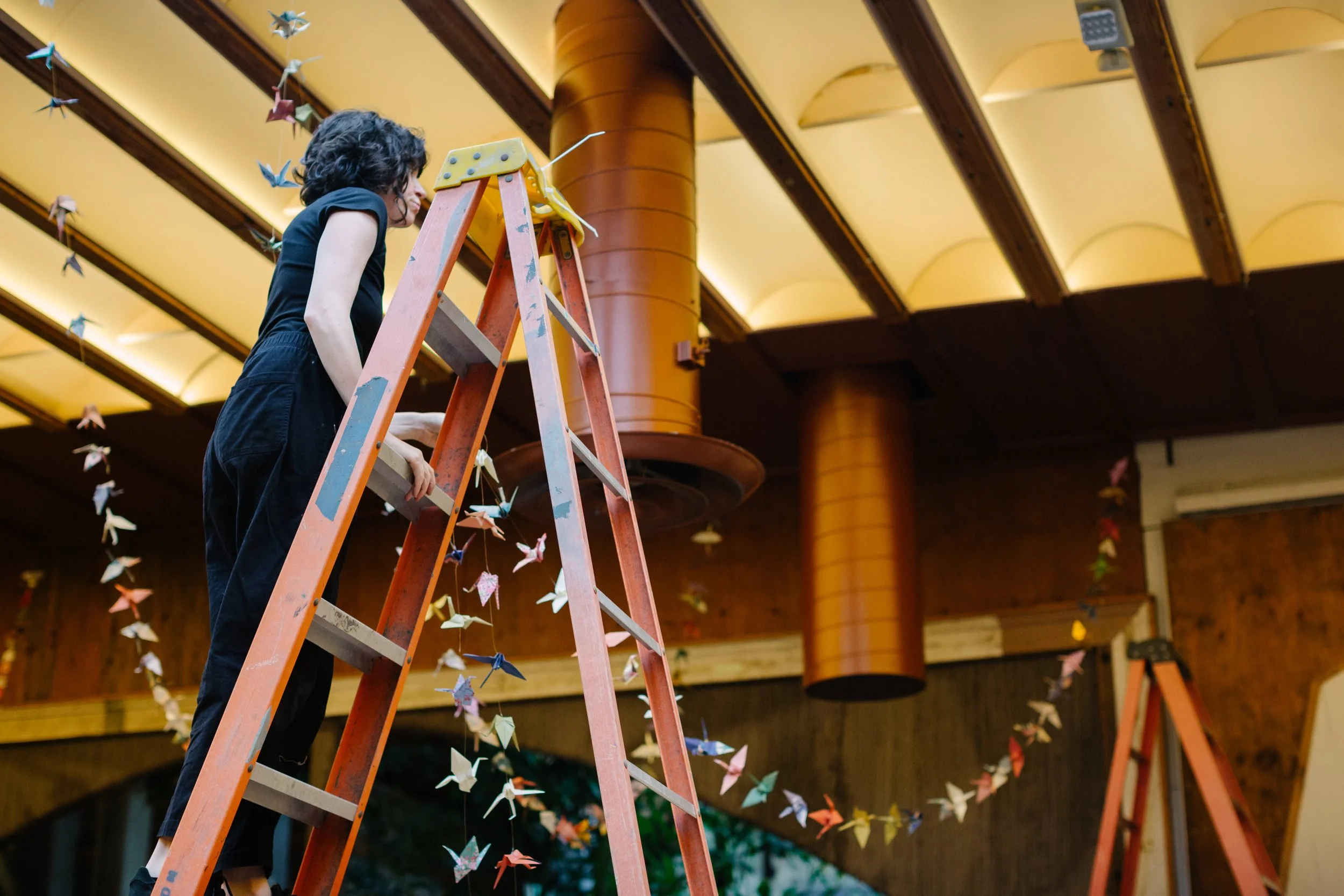A woman standing on an orange ladder hanging colorful origami paper cranes in a room with wooden walls and ceiling, decorated with strings of paper cranes.