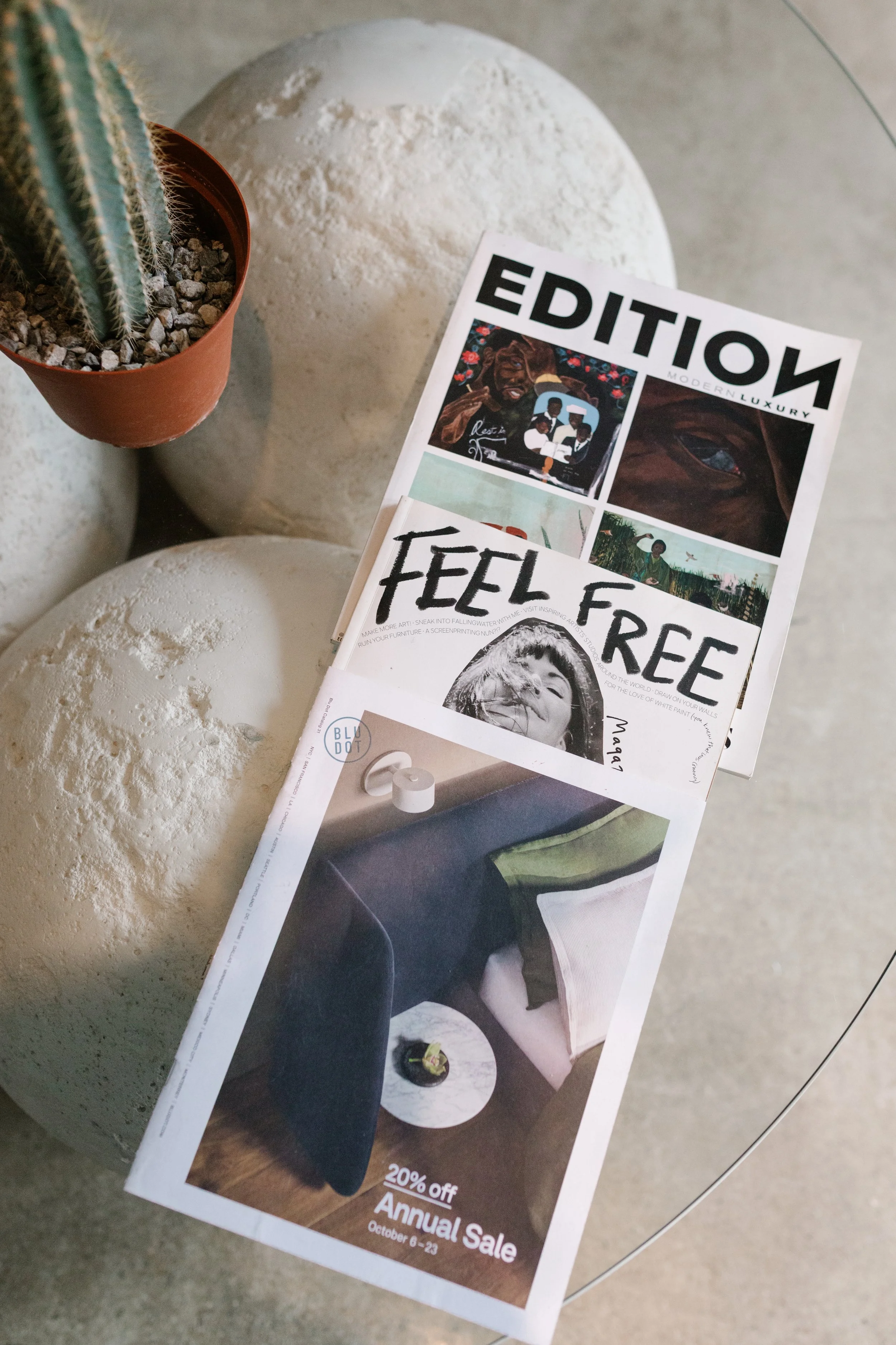 A stack of magazines and catalogs on a glass table, next to a potted cactus plant on textured white ceramic stools.