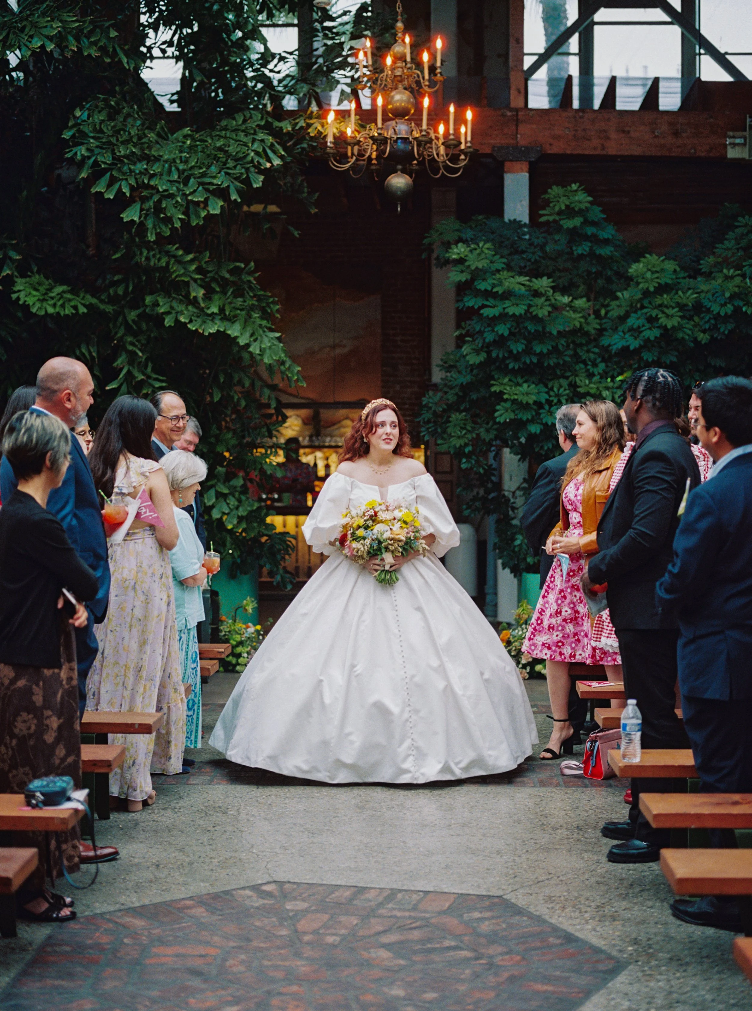 A bride in a white wedding gown holding a bouquet. Guests are standing on either side of her, dressed in formal and semi-formal attire. There are lush green trees, a chandelier with lit candles hanging above, and warm lighting in the background.