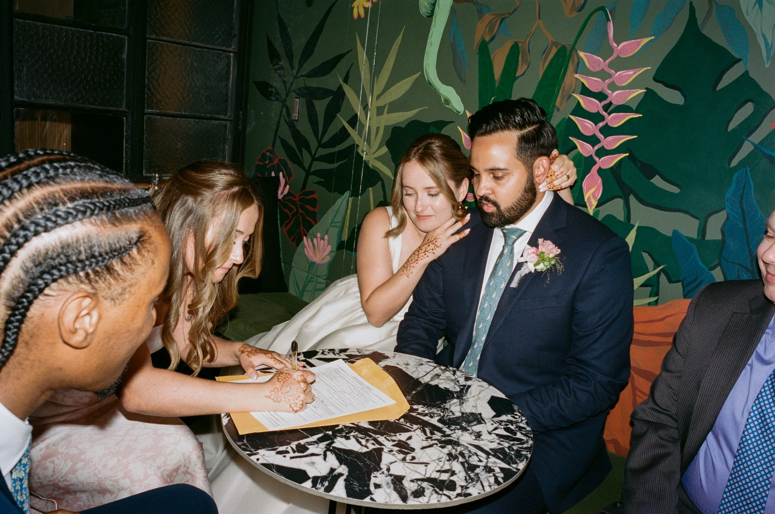 A wedding ceremony with a couple signing legal documents while sitting at a marble table, surrounded by friends. The bride is touching the groom's shoulder, with friends looking on and a colorful jungle-themed mural in the background.