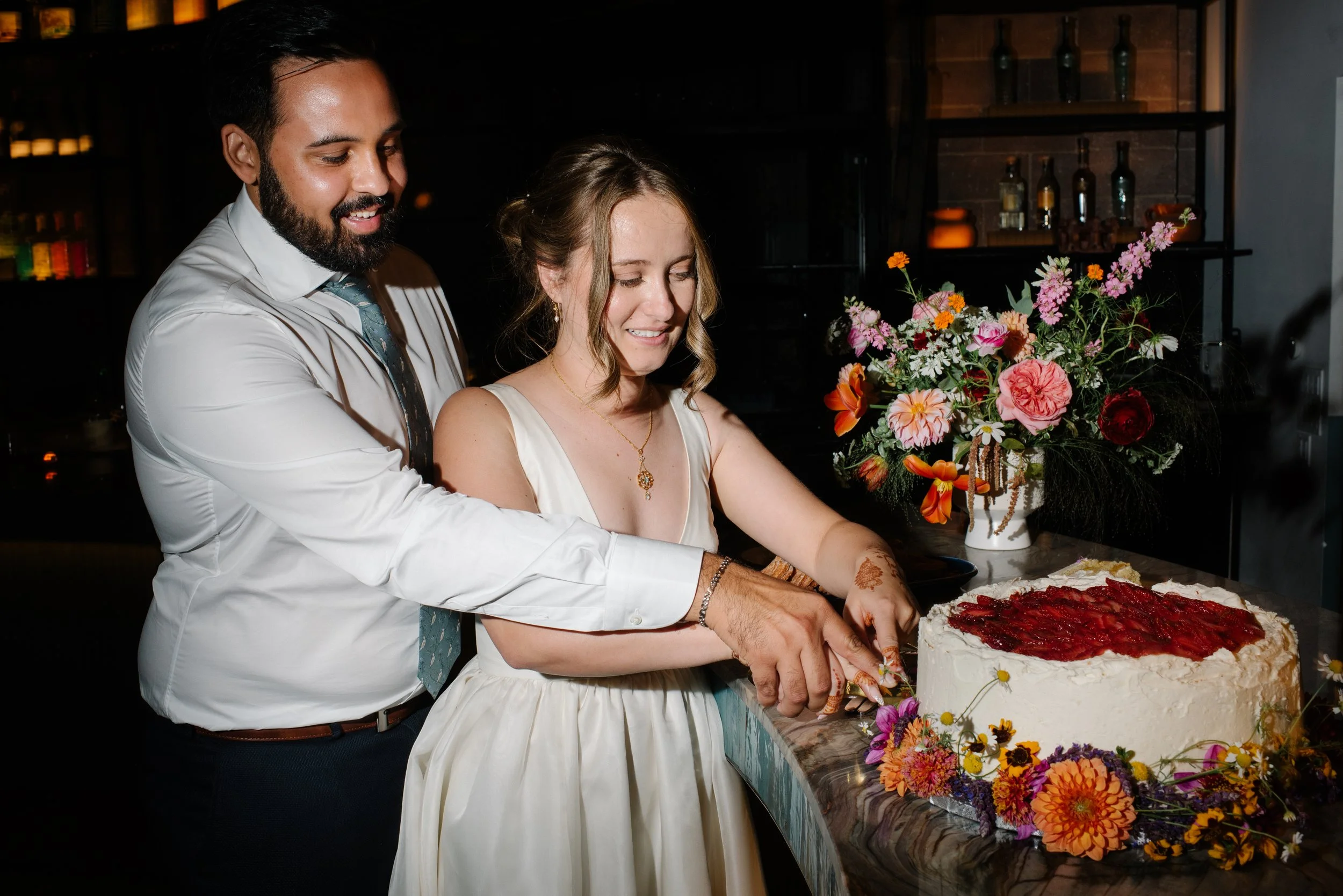 A couple cutting a wedding cake together at their celebration, with colorful flowers in a vase on the table and a dark background.