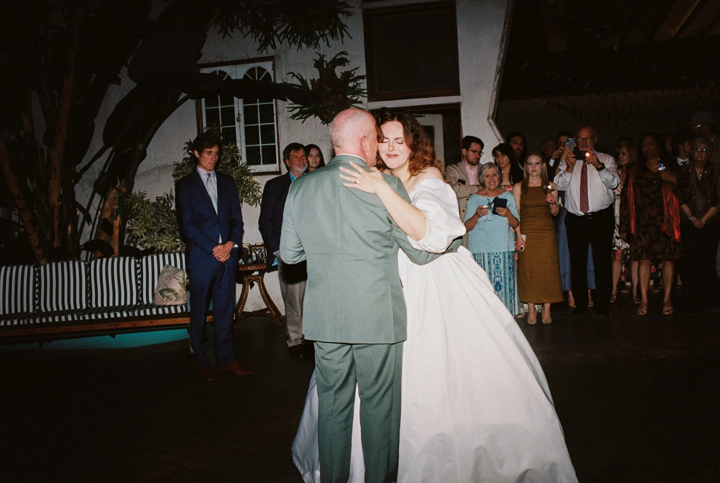 A bride and a man, possibly her father, are sharing a dance at a wedding reception. They are surrounded by guests who are watching and taking photos in an indoor venue with greenery and decorative elements.