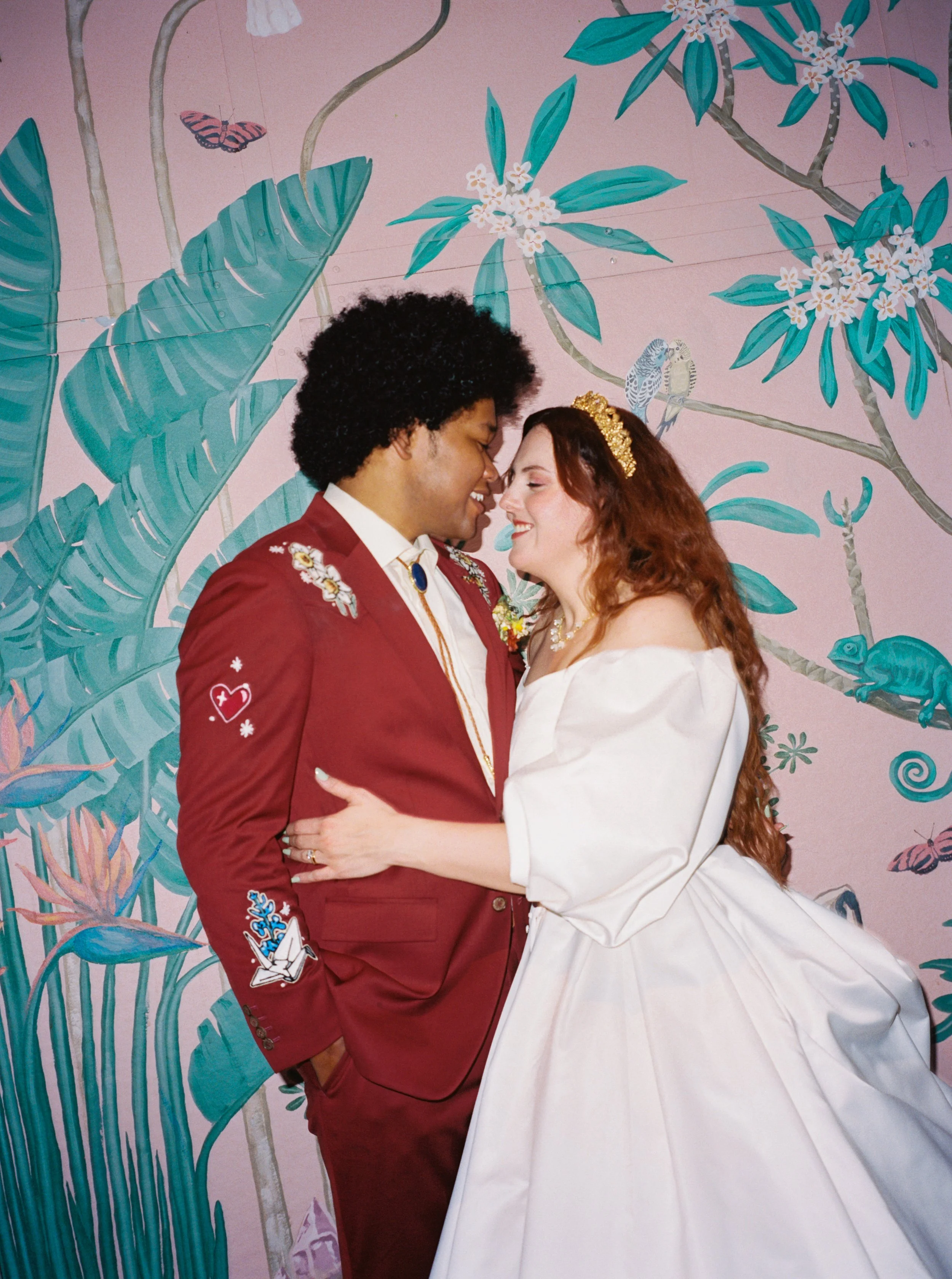A couple is standing close together, smiling and about to kiss. The man has curly black hair and is wearing a red jacket with embroidered patches, while the woman has long red hair, a gold tiara, and a white dress. The background features a colorful,