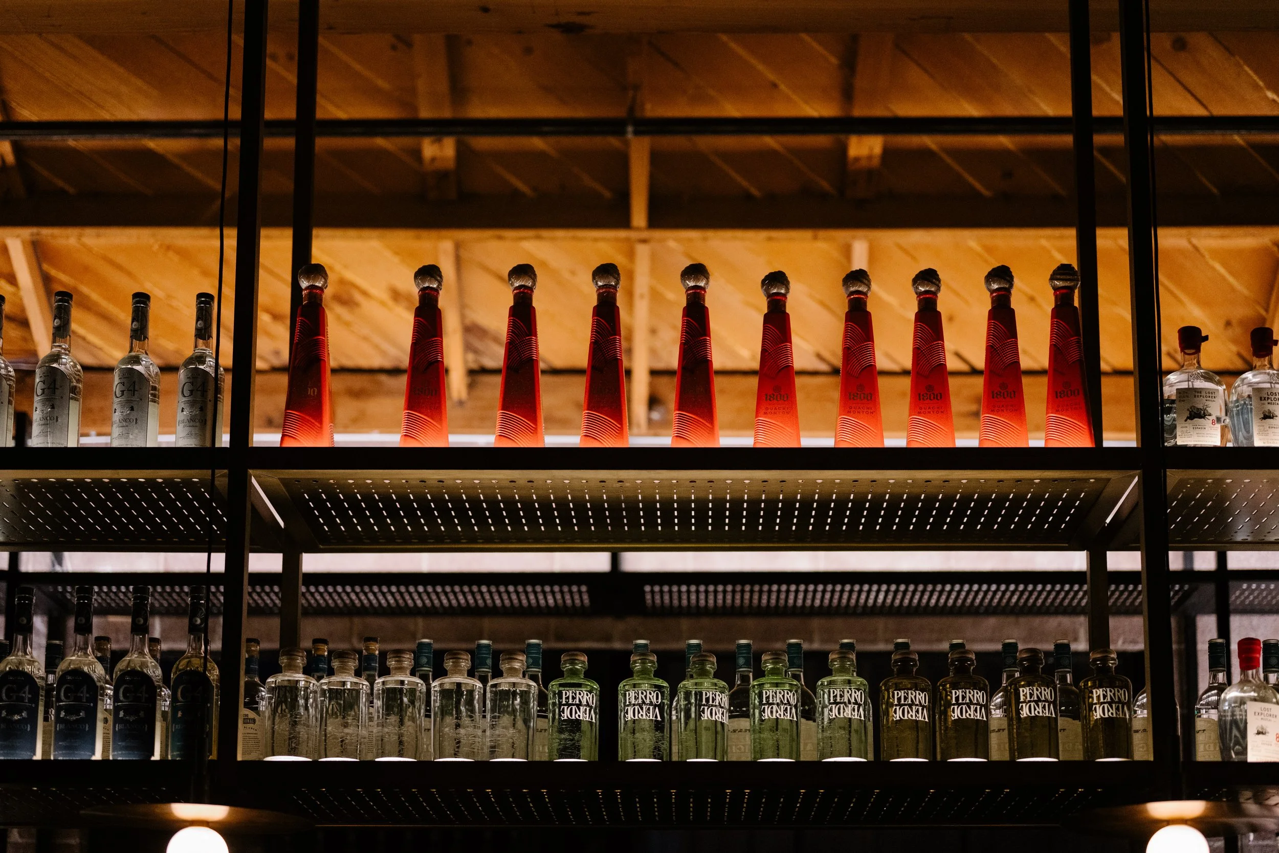 Shelves with various bottles of alcohol, including tall red bottles on the top shelf and green bottled drinks on the bottom shelf, under a wooden ceiling.