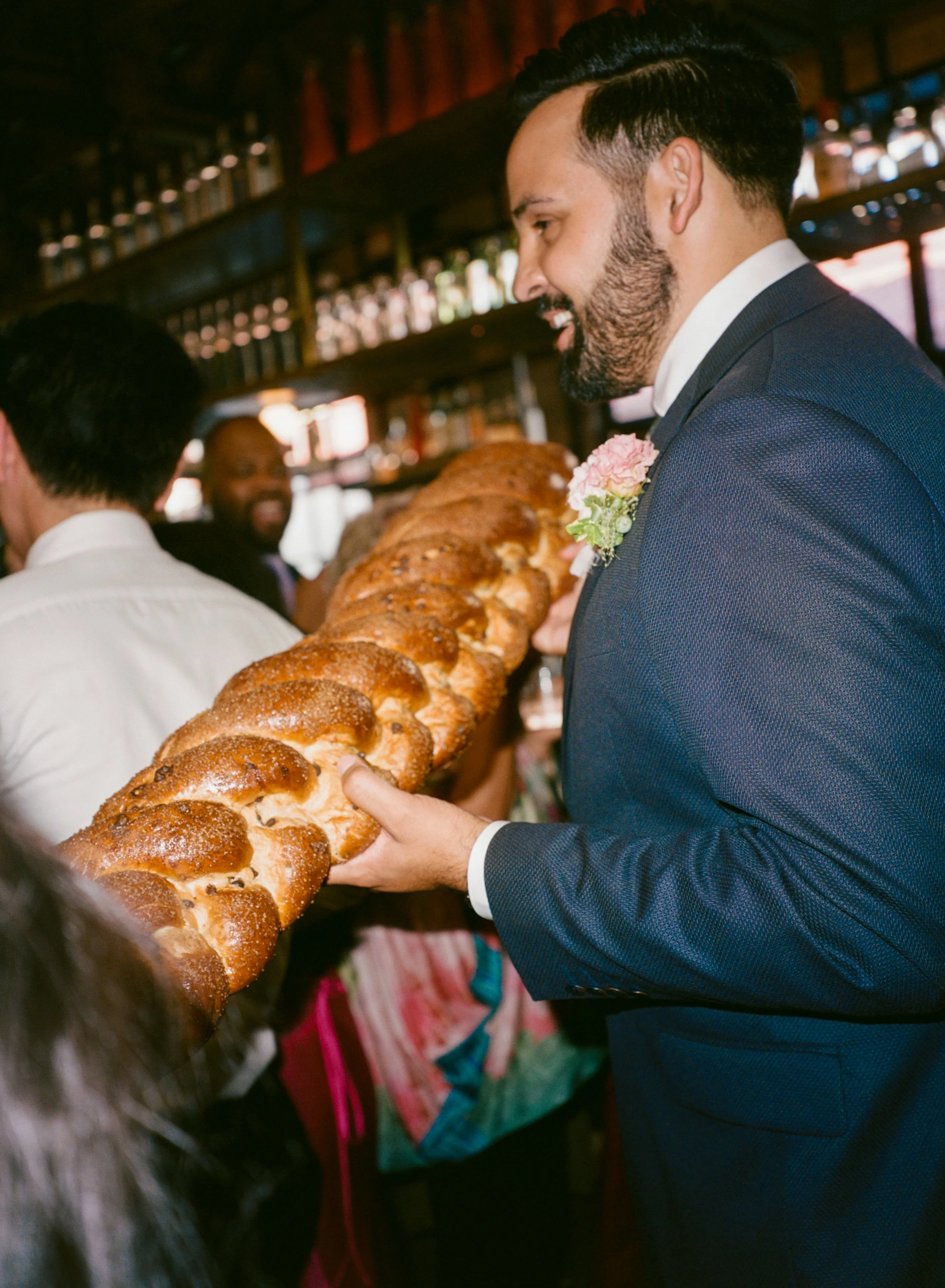 A man in a navy suit with a pink flower boutonniere holds a large braided challah bread at a Jewish wedding with others in the background and bottles on shelves behind them.