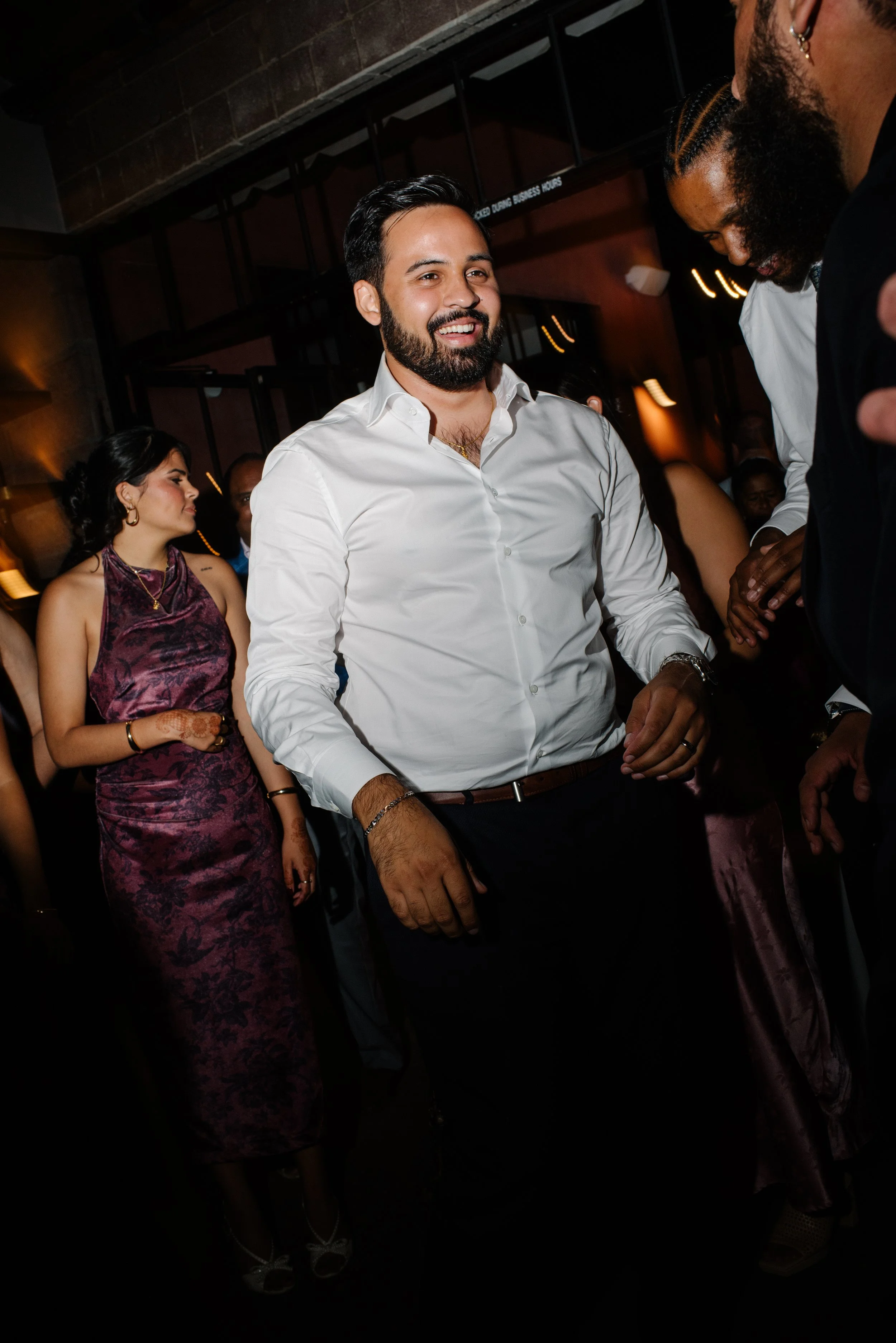 A groom in a white shirt smiling and talking to others, with a woman in a purple dress standing nearby, inside a dimly lit venue.