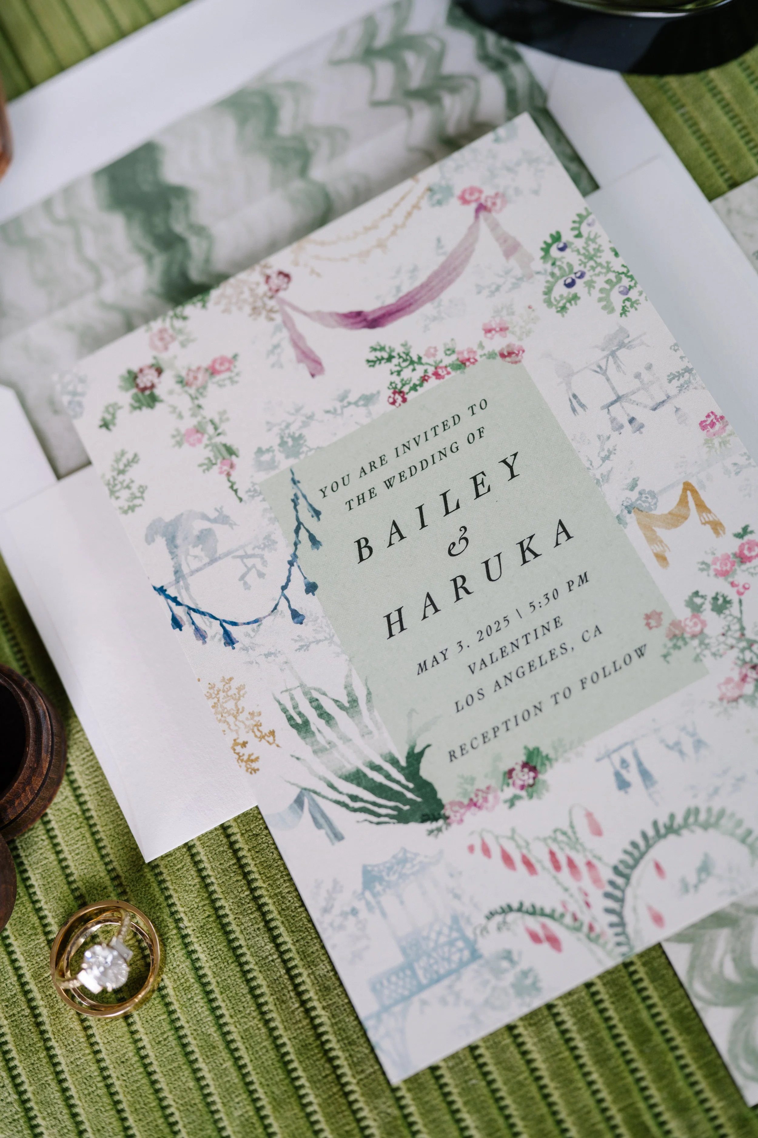 Wedding invitation with floral illustrations, announcing the wedding of Bailey and Haruka on May 3, 2025, in Los Angeles, CA, against a green surface.