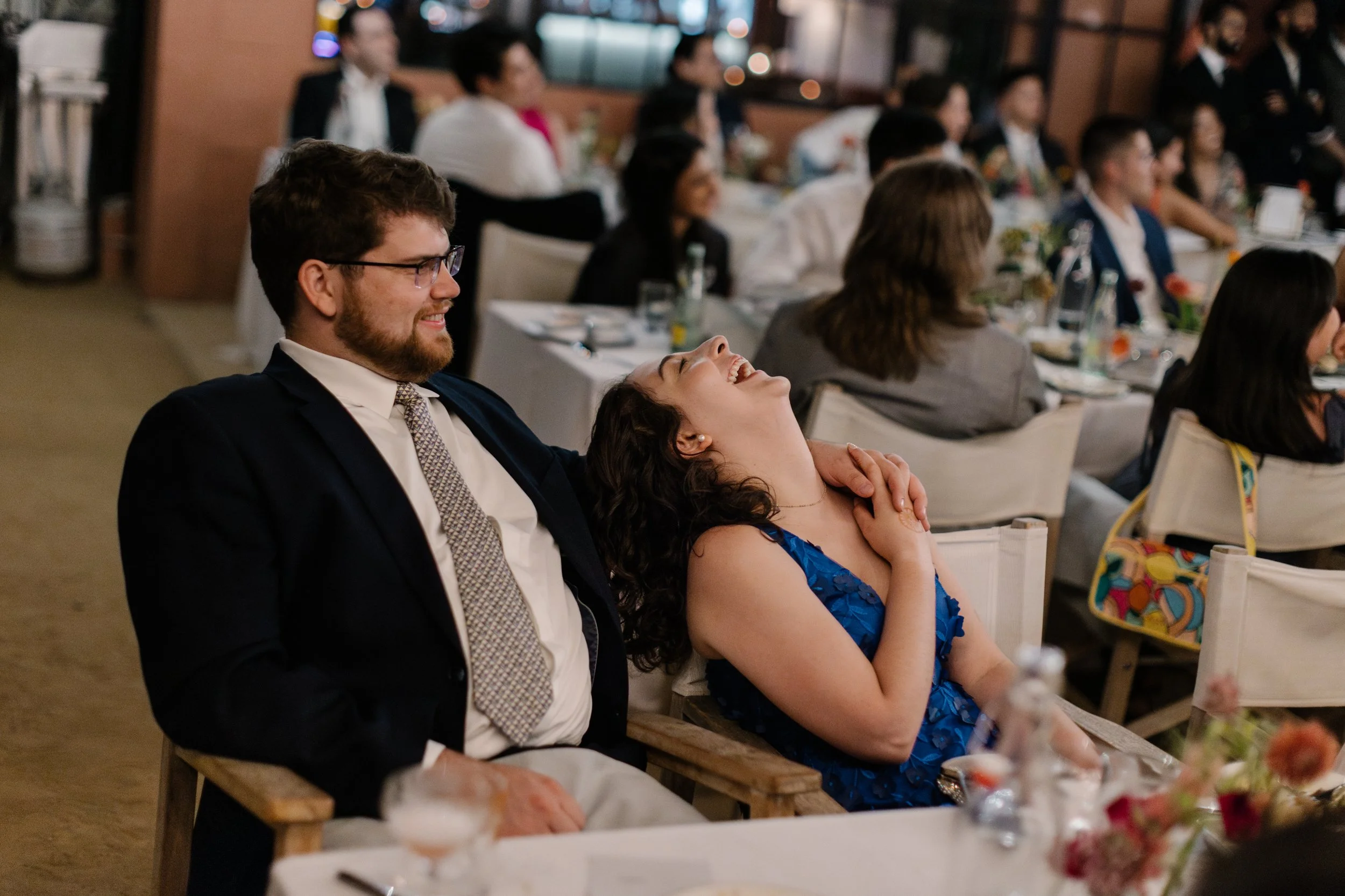 A man and a woman are sitting at a table, laughing and enjoying a moment together at a wedding with other guests seated around tables in the background. The woman is throwing her head back in laughter.