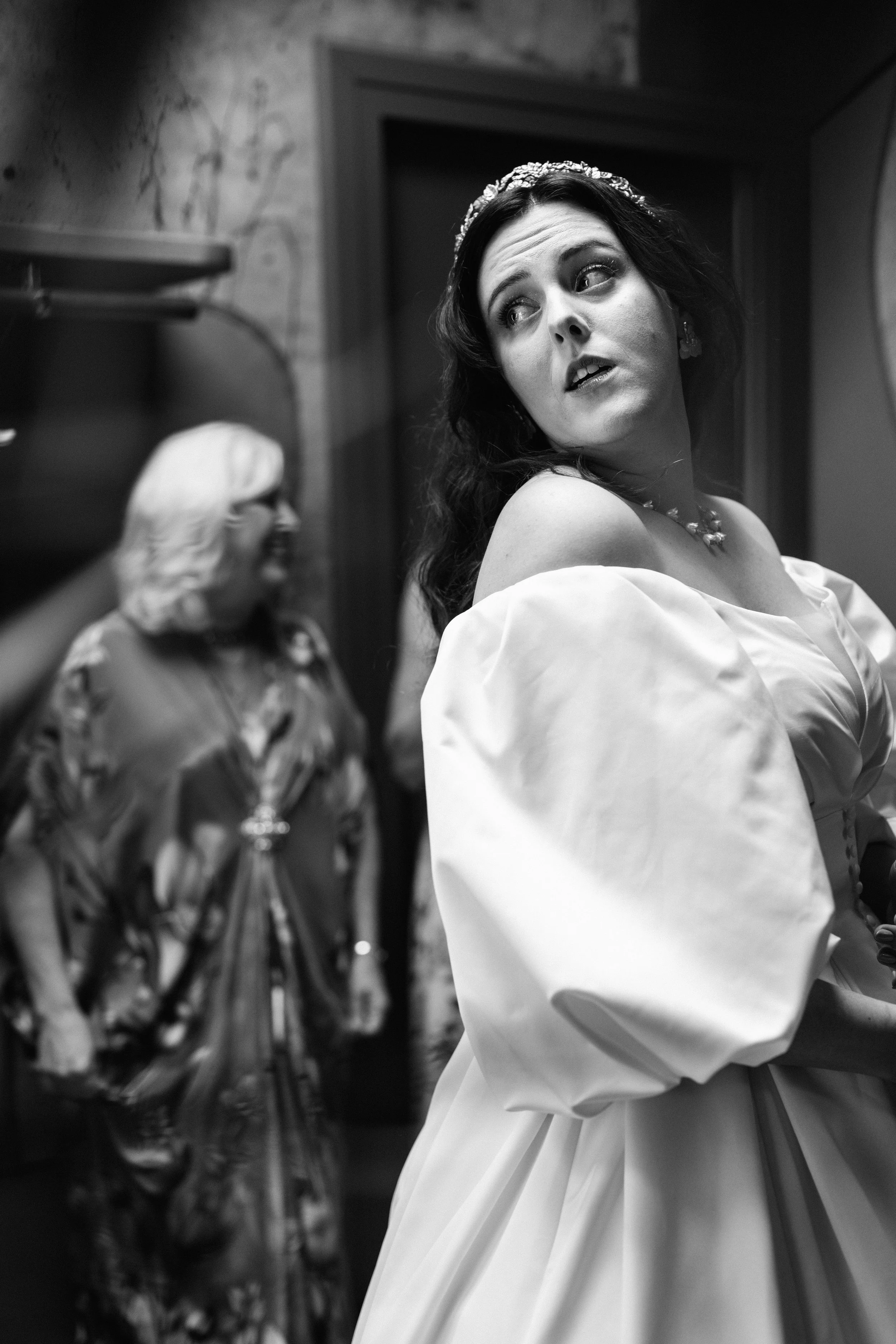 A bride in a wedding dress with puffed sleeves, wearing a tiara and jewelry, looking at the mirror in a room with older woman in the background.
