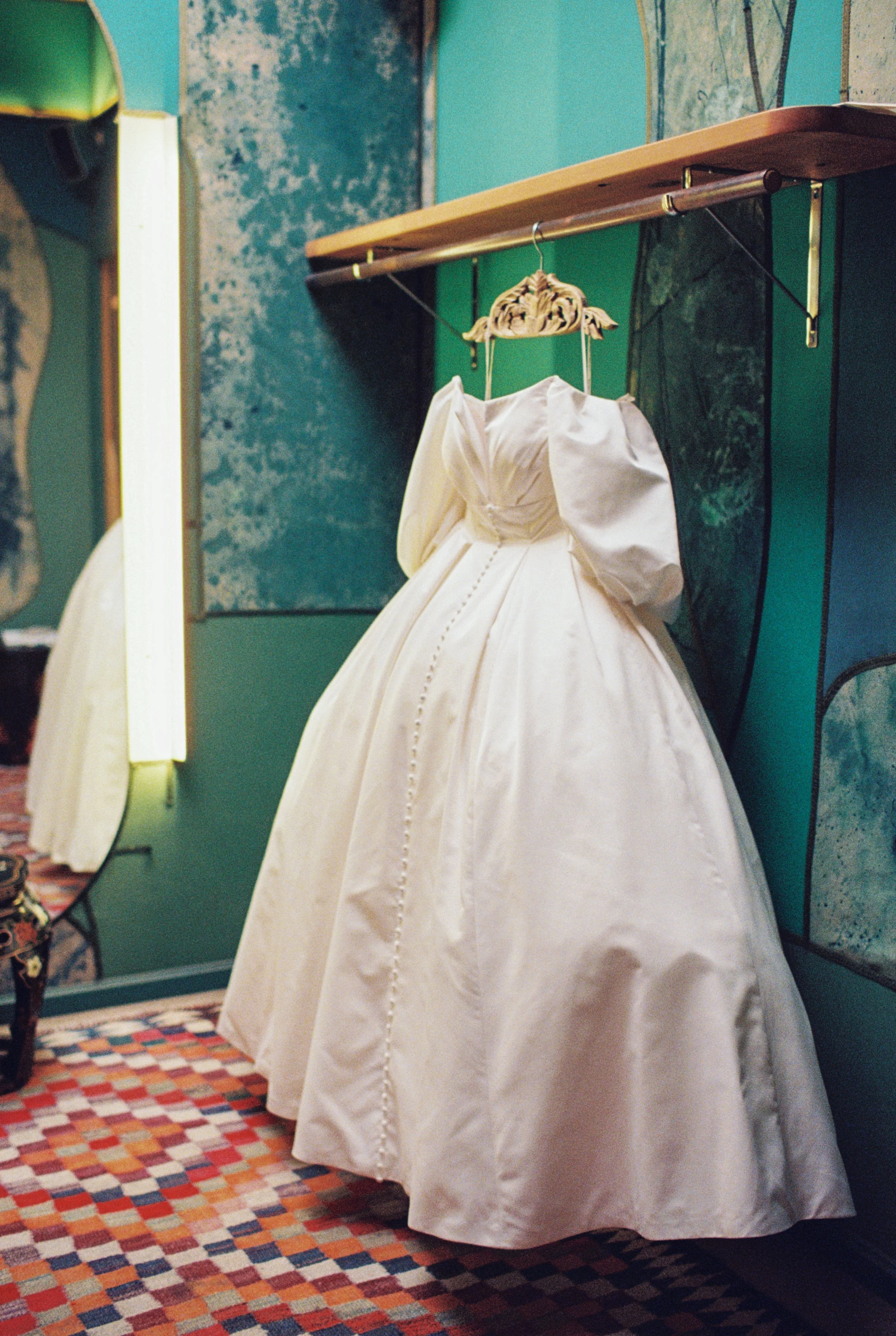 A white wedding dress hanging on a decorative hanger in front of a colorful wall. The dress is displayed in front of a mirror and the room has a patterned carpet.