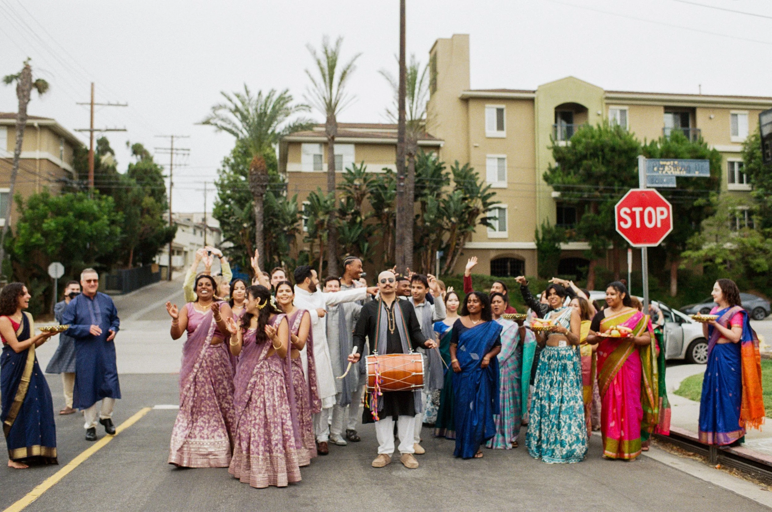 A group of people dressed in traditional Indian clothing participating in an Indian wedding baraat on a city street, with some holding bowls and others playing musical instruments, and a stop sign visible in the background.