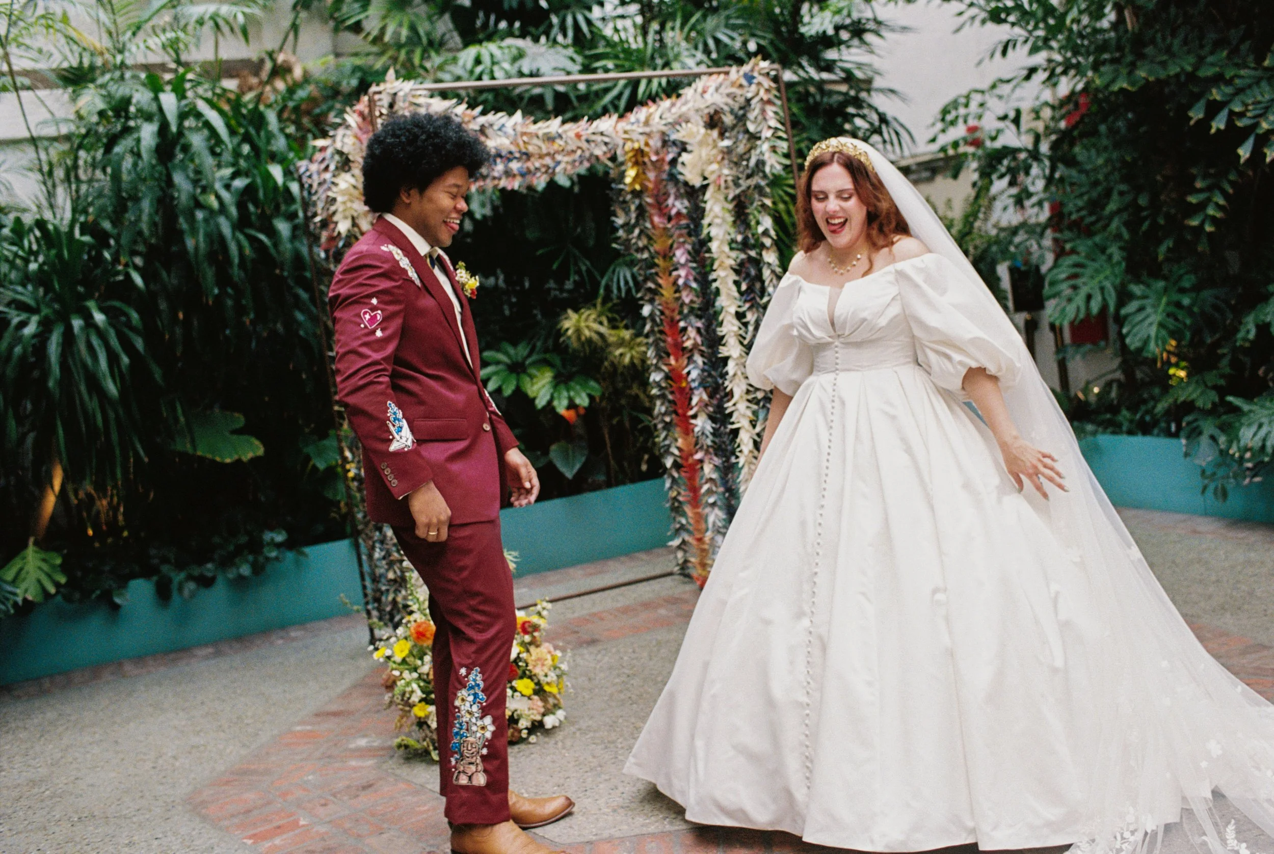A bride with long red hair in a white gown dancing with a groom wearing a burgundy suit at a wedding reception surrounded by green plants and floral decorations.