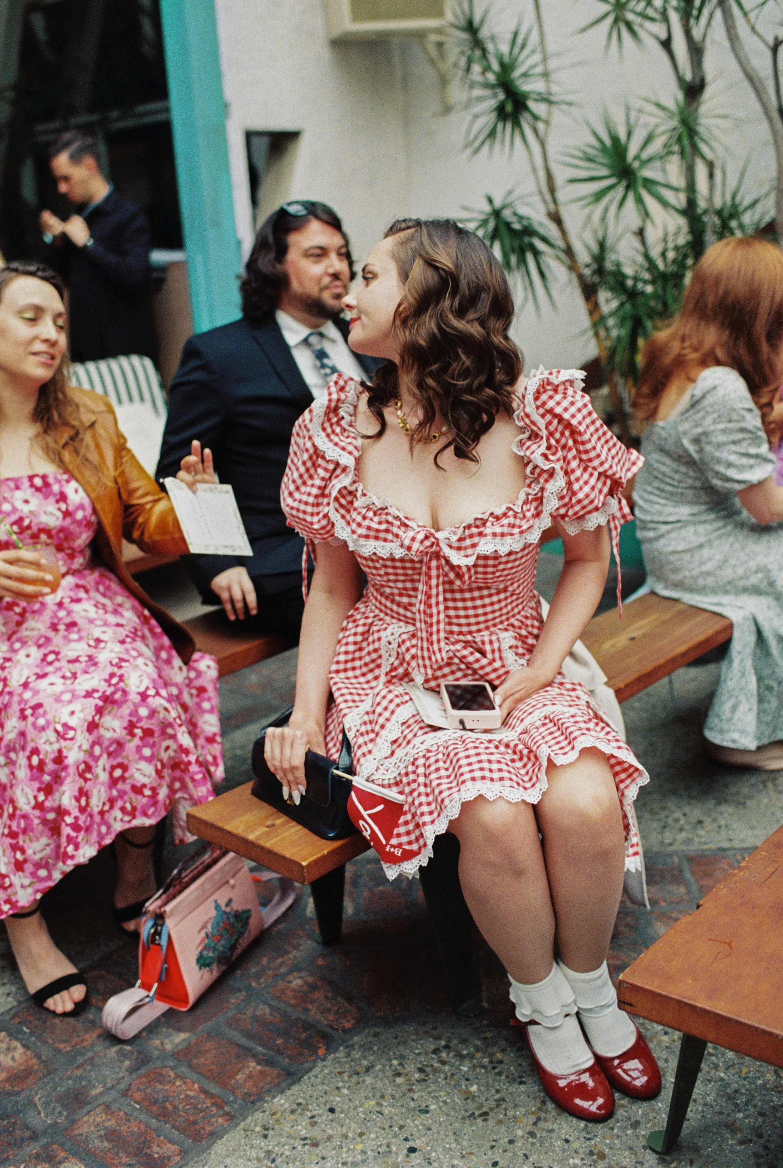 A woman in a red and white checkered dress with ruffled sleeves sitting on a bench, holding a phone and wearing vintage-style shoes, at a wedding with other people dressed in colorful outfits and plants in the background.