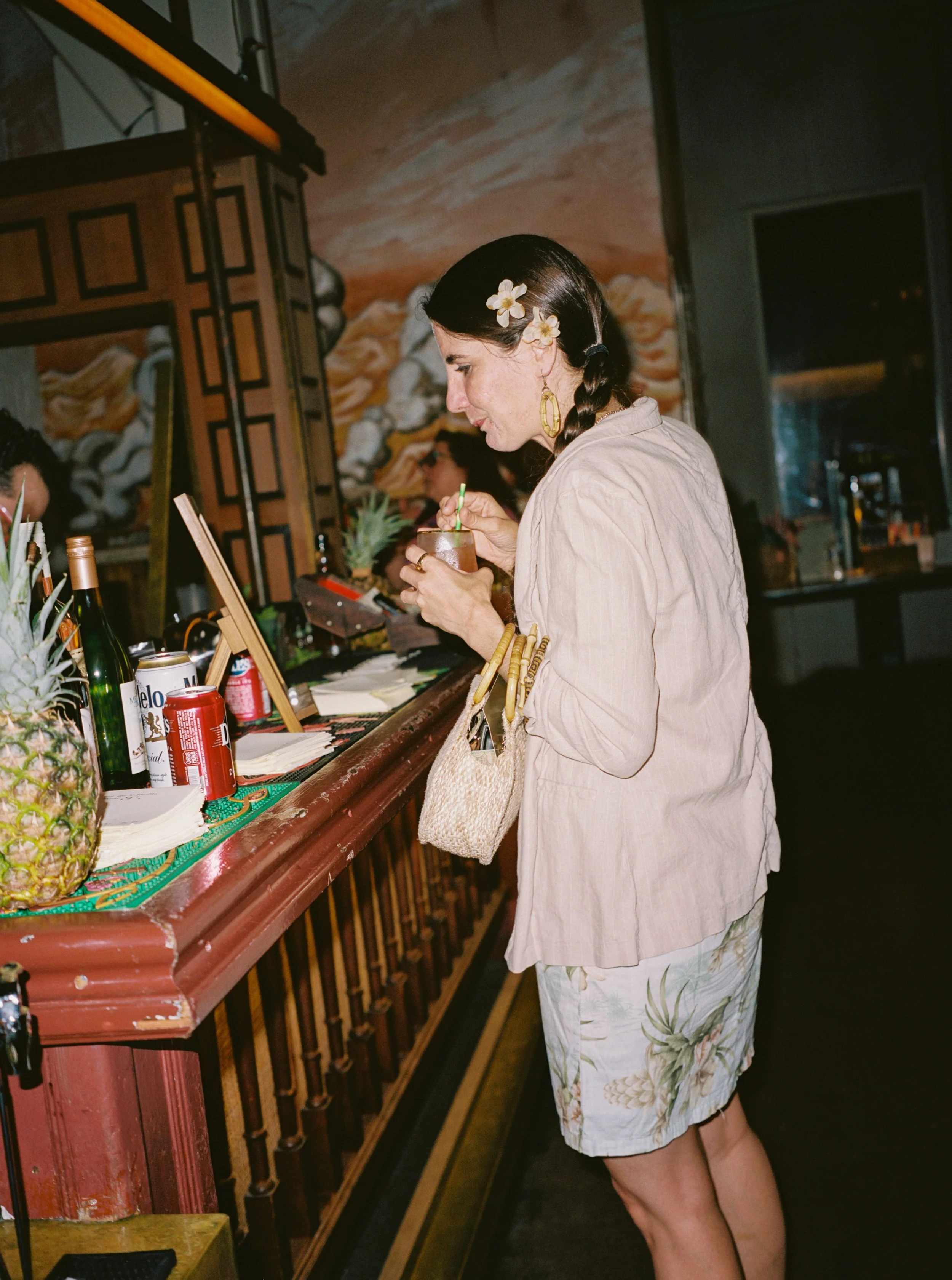 A woman with floral earrings and a flower in her hair, holding a drink with a straw, standing at a bar with various bottles and snacks, including a pineapple and cans, in a restaurant with a mural painting in the background.