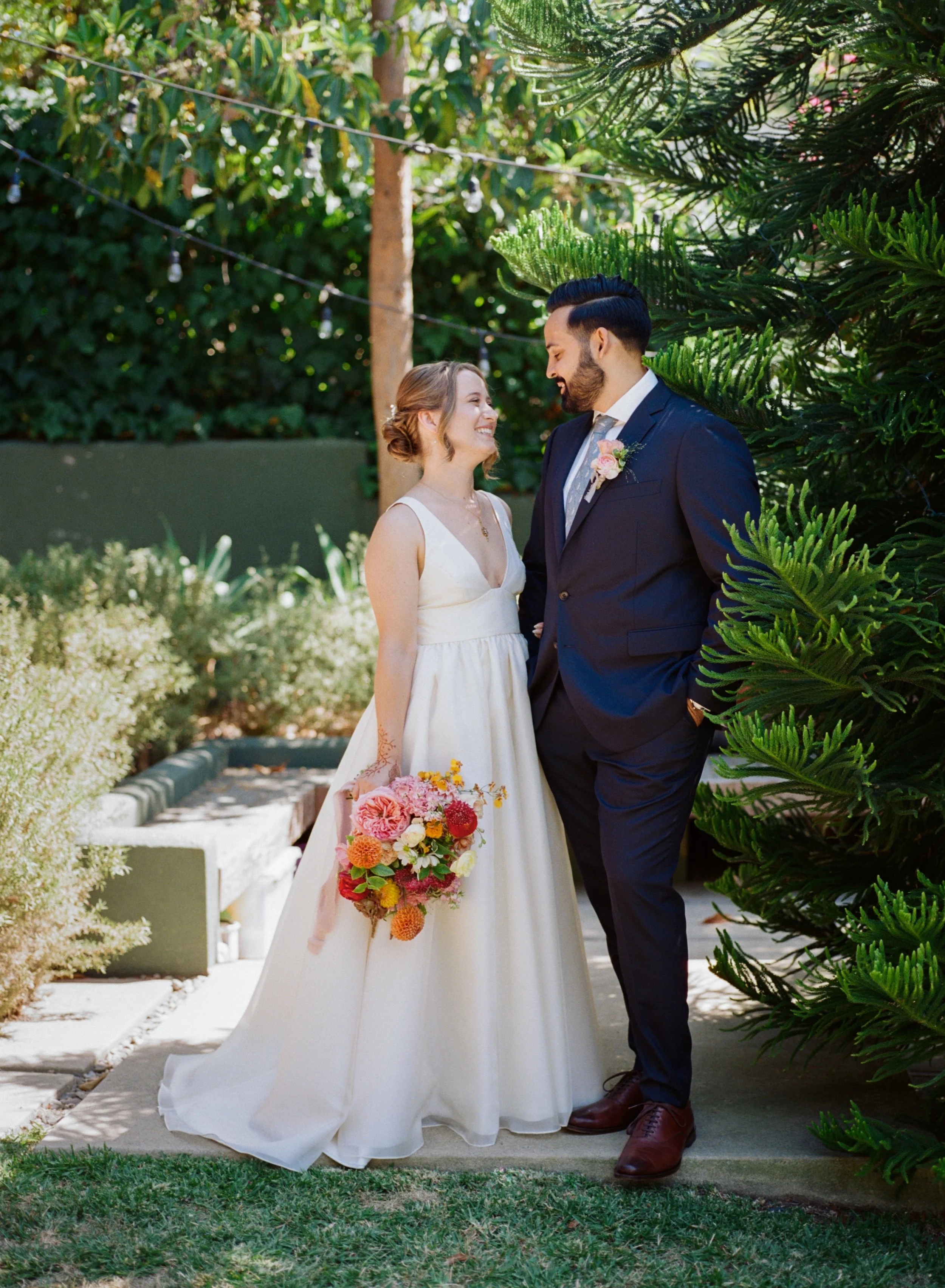 A bride and groom in wedding attire standing outdoors, smiling at each other, surrounded by lush green plants and trees, with sunlight filtering through the leaves.