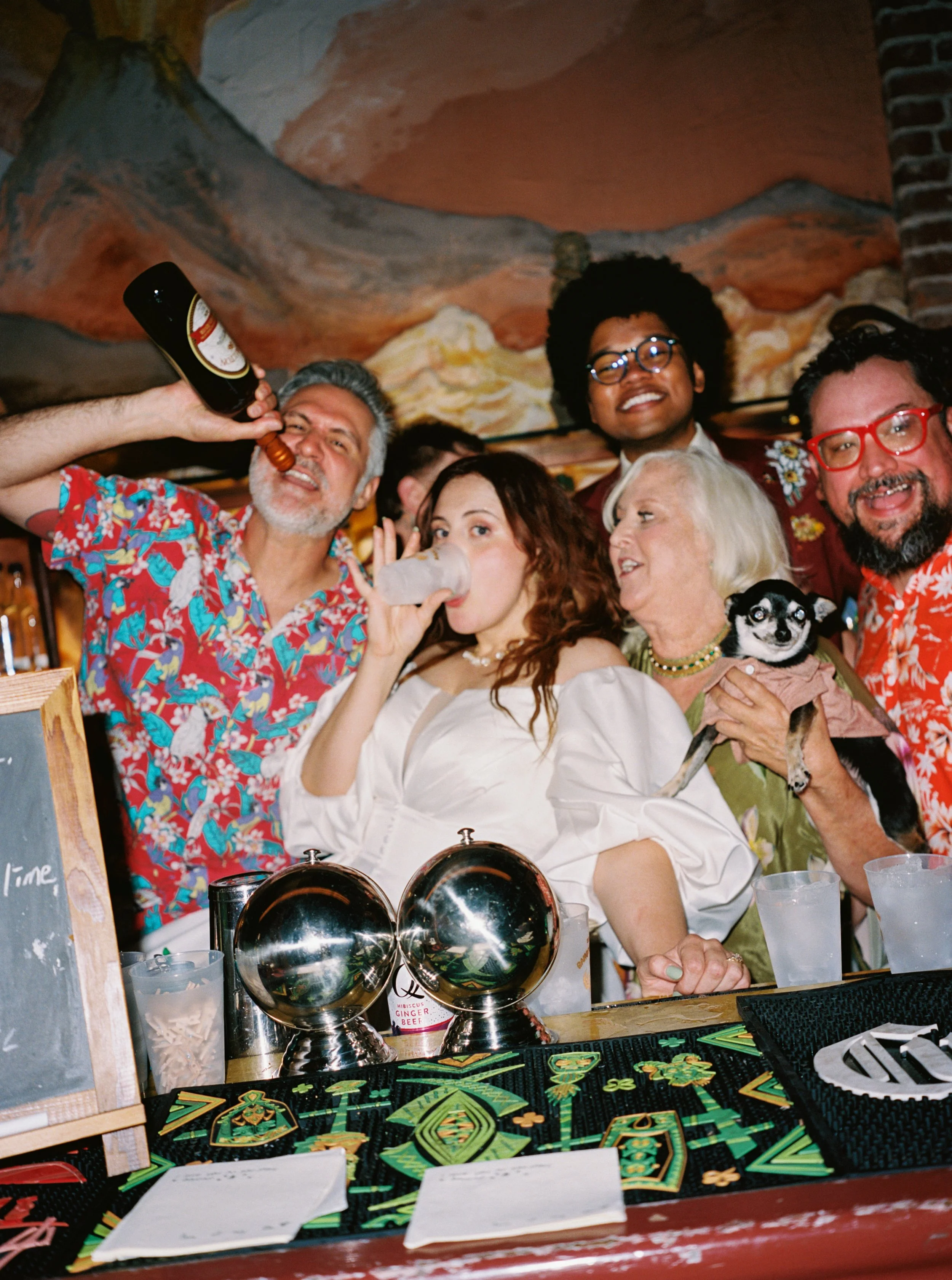 Group of diverse people celebrating with drinks at a bar, some smiling and chatting, with a small dog being held, and colorful decorations on the table.