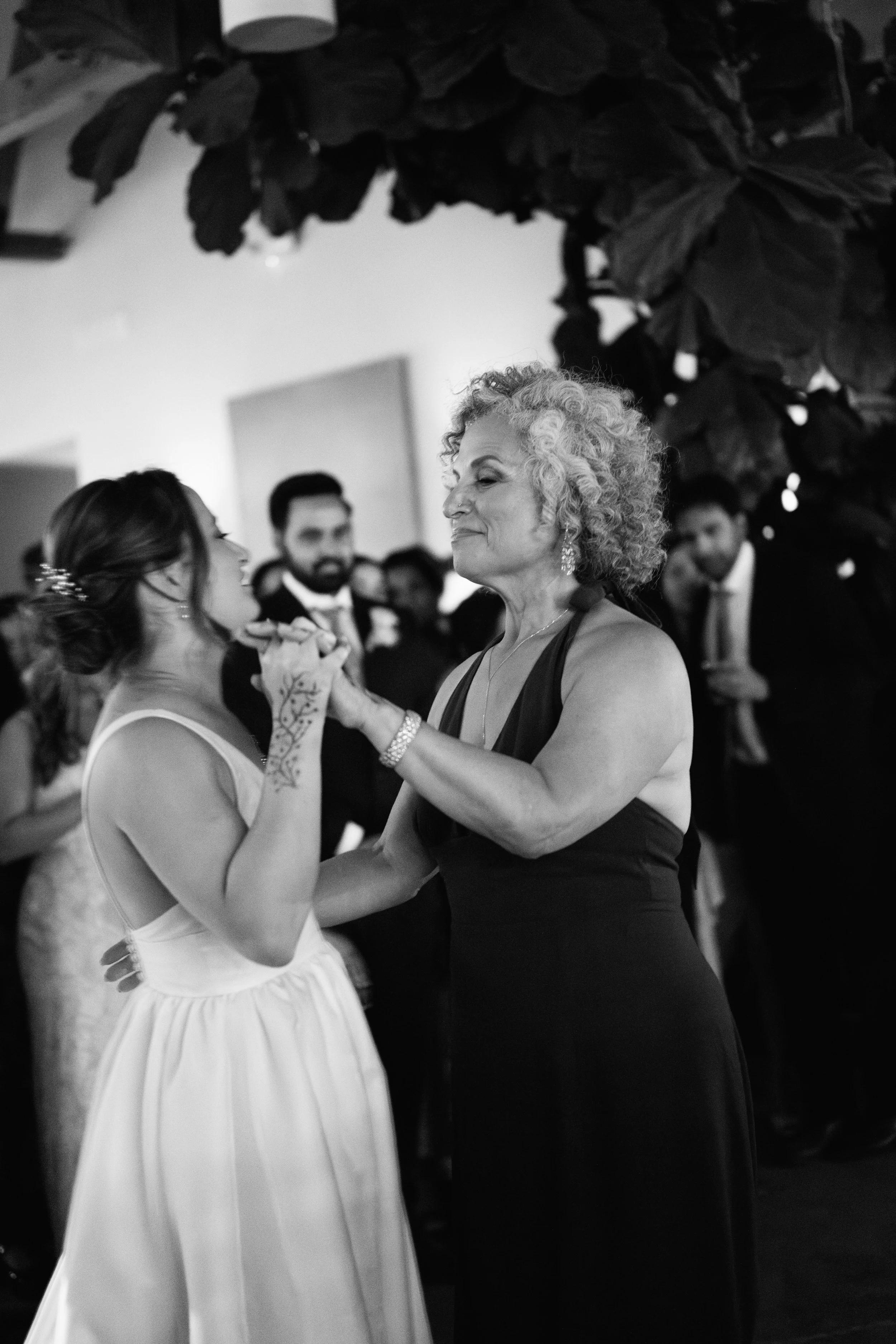 A bride dances with an older woman, possibly her mother, at a wedding reception, with guests in the background.