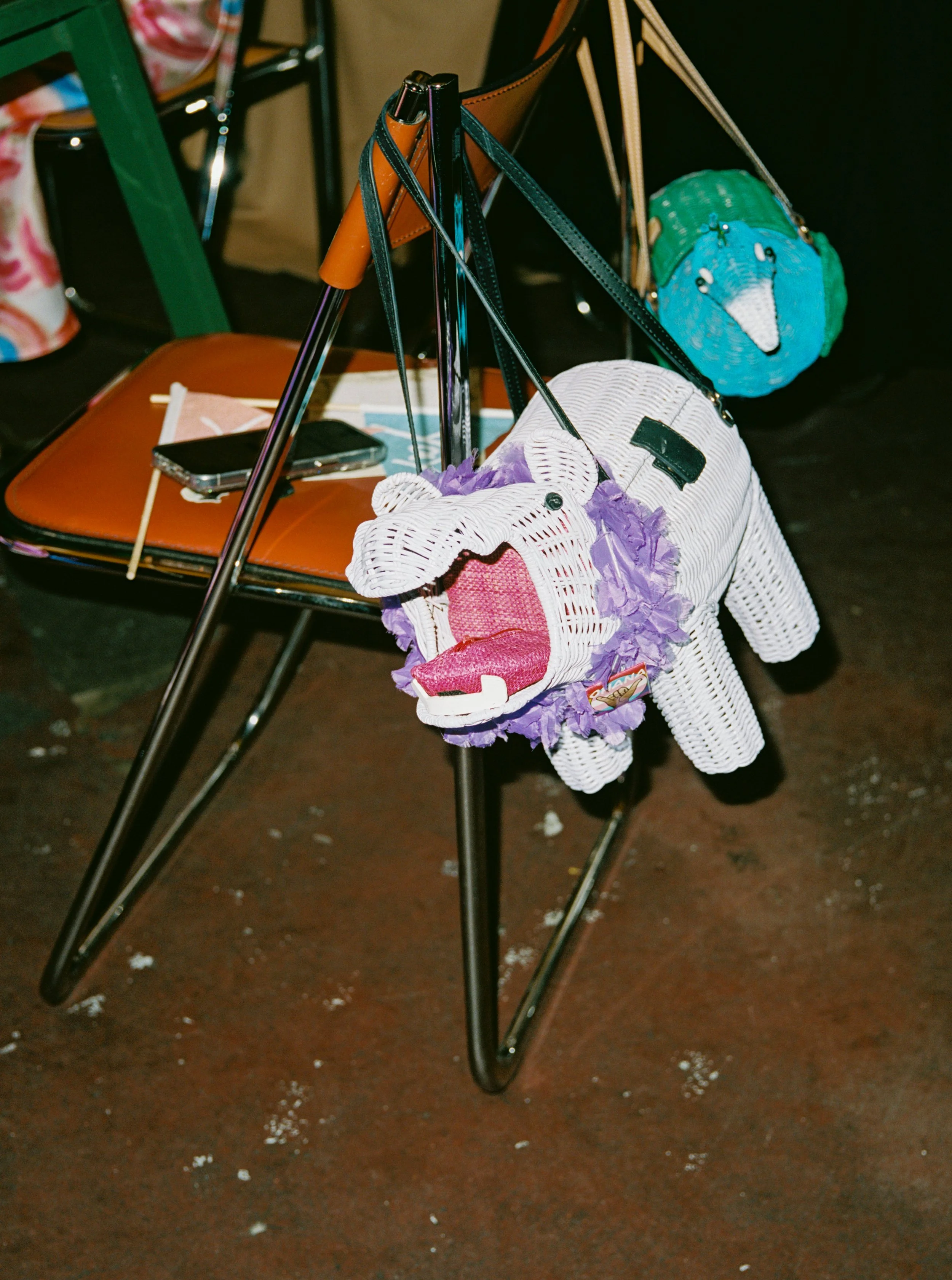A purple wicker purse shaped like a hippo, hanging from a black chair. In the background, there's a turquoise handbag shaped like a peacock also hanging from the chair.