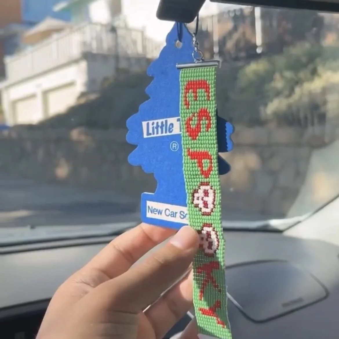 A person's hand holding car air freshener hanging from the rearview mirror inside a vehicle, with a background of a street scene.