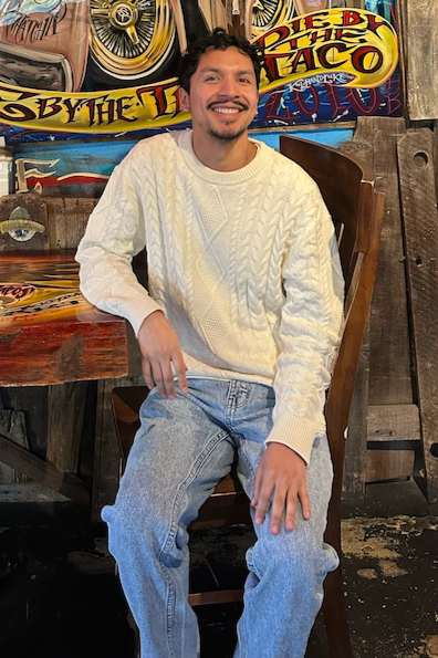 A man sitting on a wooden chair, smiling, wearing a white cable-knit sweater and light jeans, in a rustic setting with skateboard art on the wall behind him.