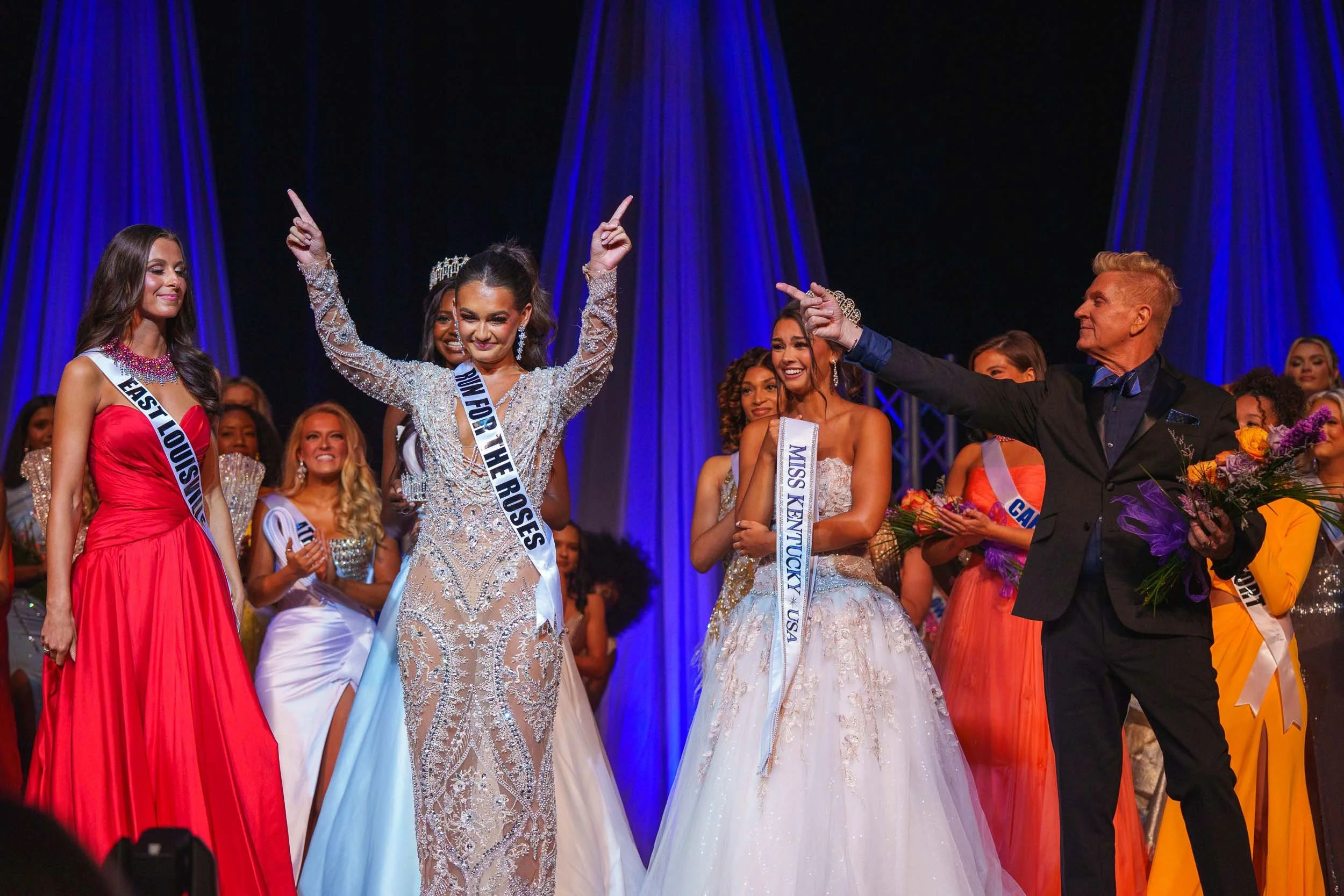 Beauty pageant winner wearing a sash and crown, smiling, surrounded by other contestants, on stage with blue curtains in the background.