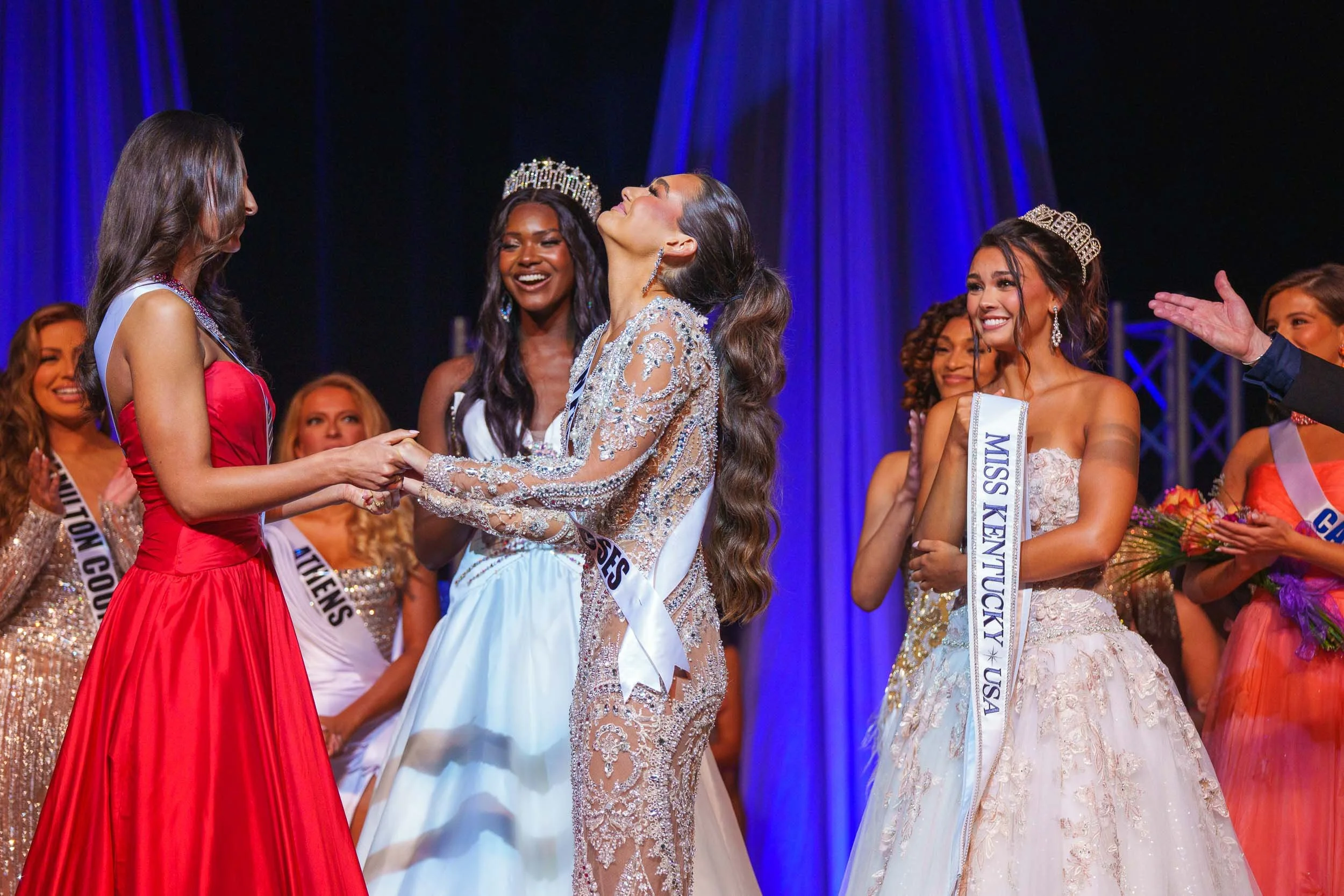Miss Kentucky is crowned during a beauty pageant, wearing a crown and a sash, with other contestants and an audience in the background.