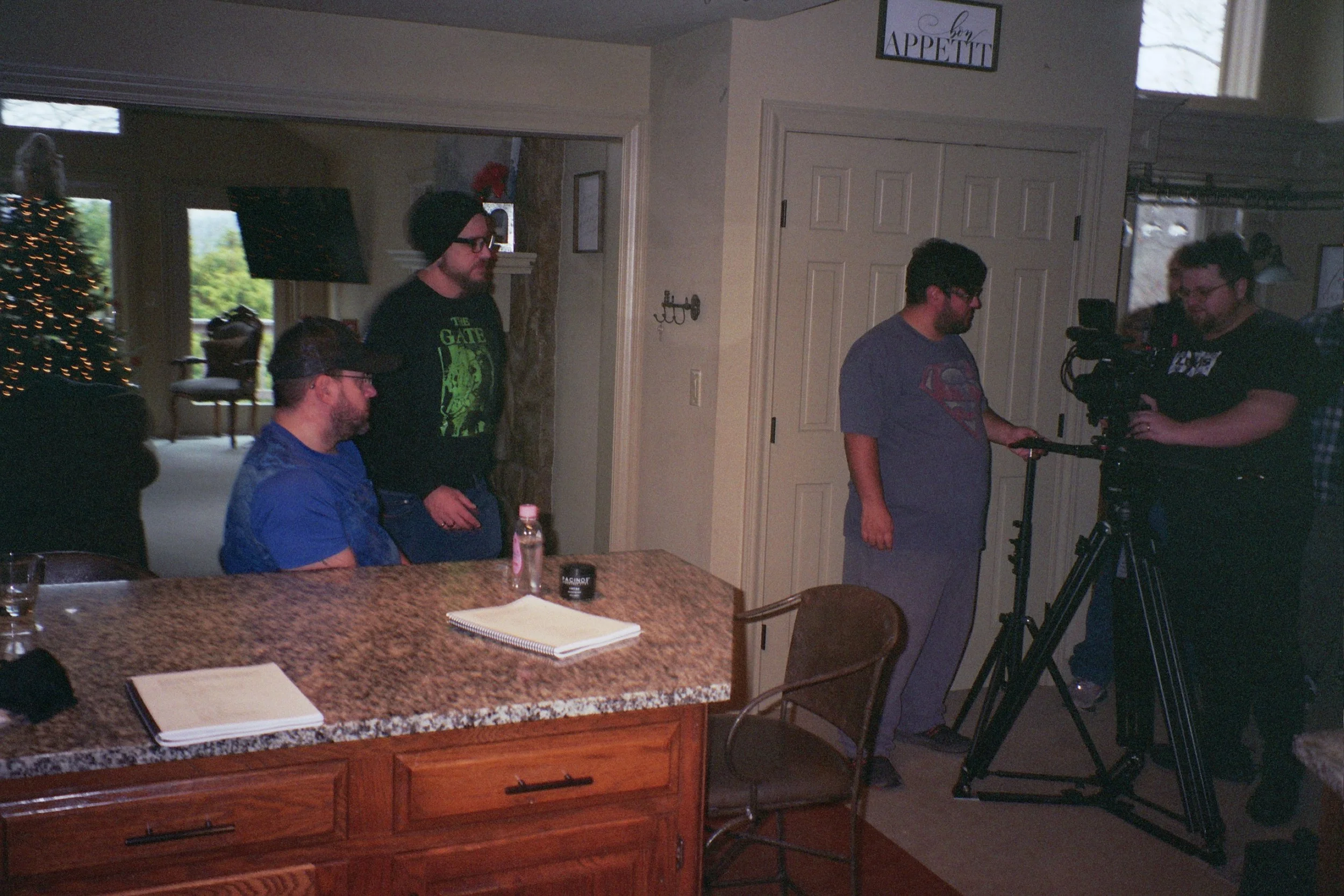 People filming a video in a room with a sign that reads 'Bon Appetit' on the wall, Christmas tree visible in the background, and a granite kitchen counter with notebooks and a water bottle in the foreground.