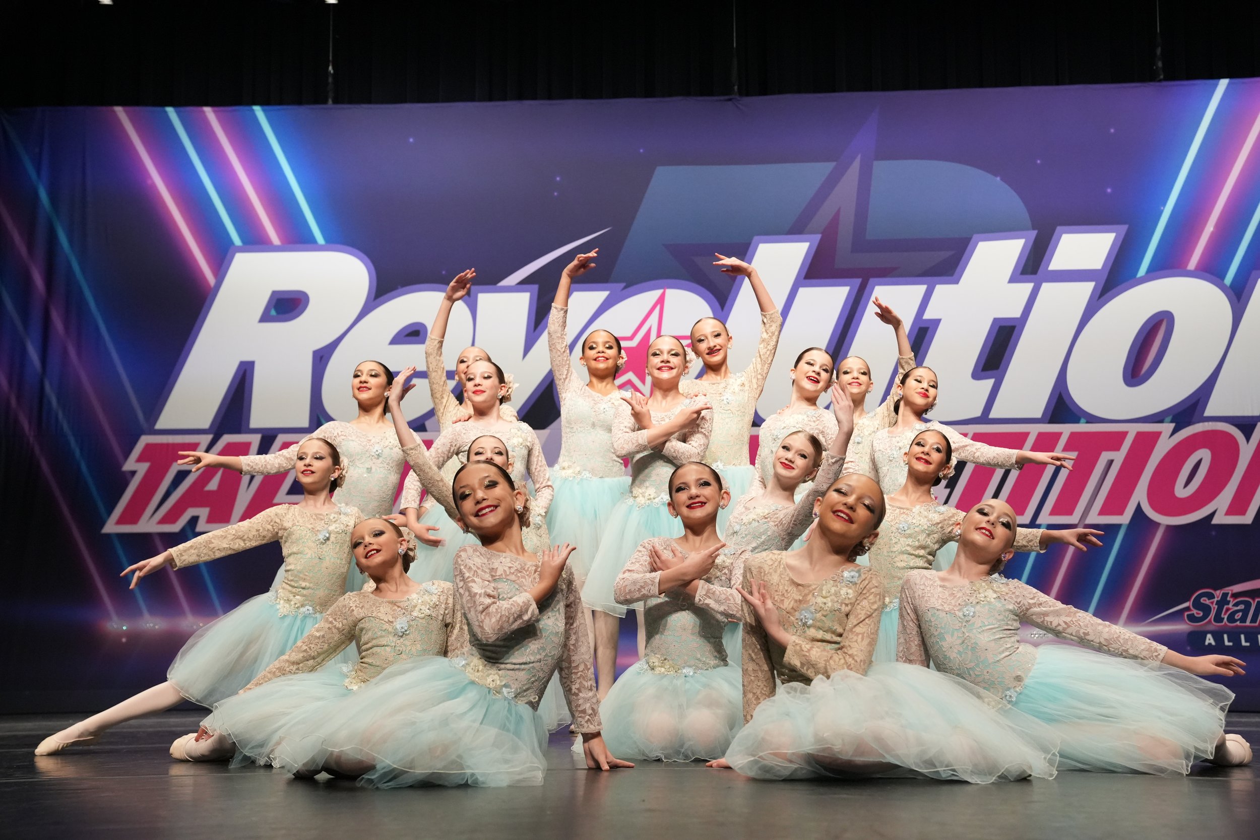 Group of young ballet dancers performing on stage in costume at a competition with a colorful backdrop.