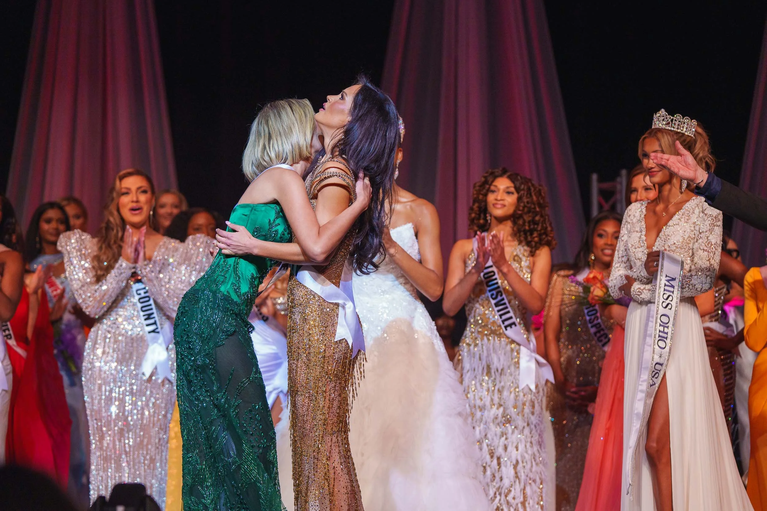 A beauty pageant scene with women in evening gowns on stage, two women hug in the foreground, one wearing a sash and crown, surrounded by other contestants clapping.