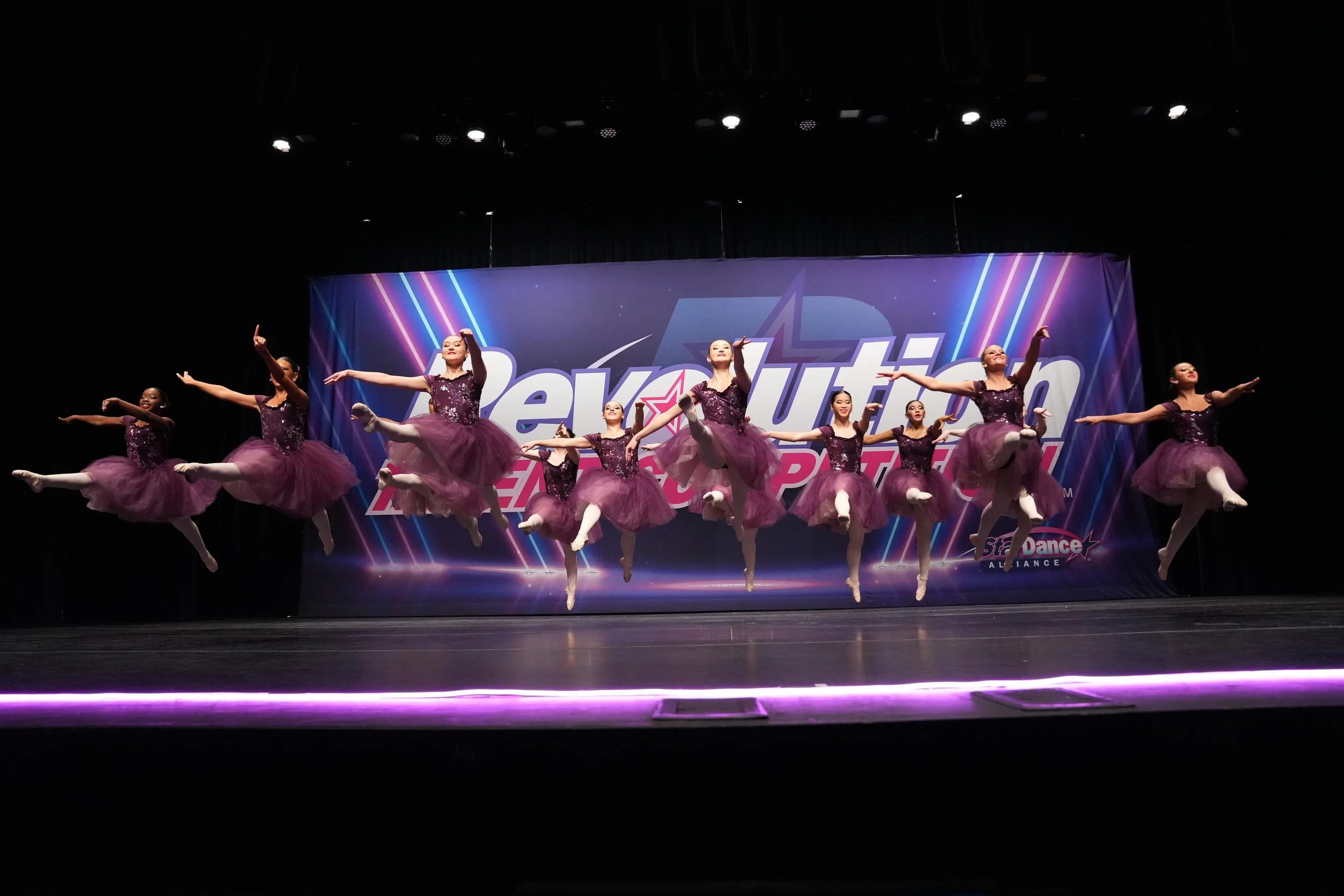 Ballet dancers in purple tutus performing a jump on stage at a dance competition with a "Revolution" themed backdrop.