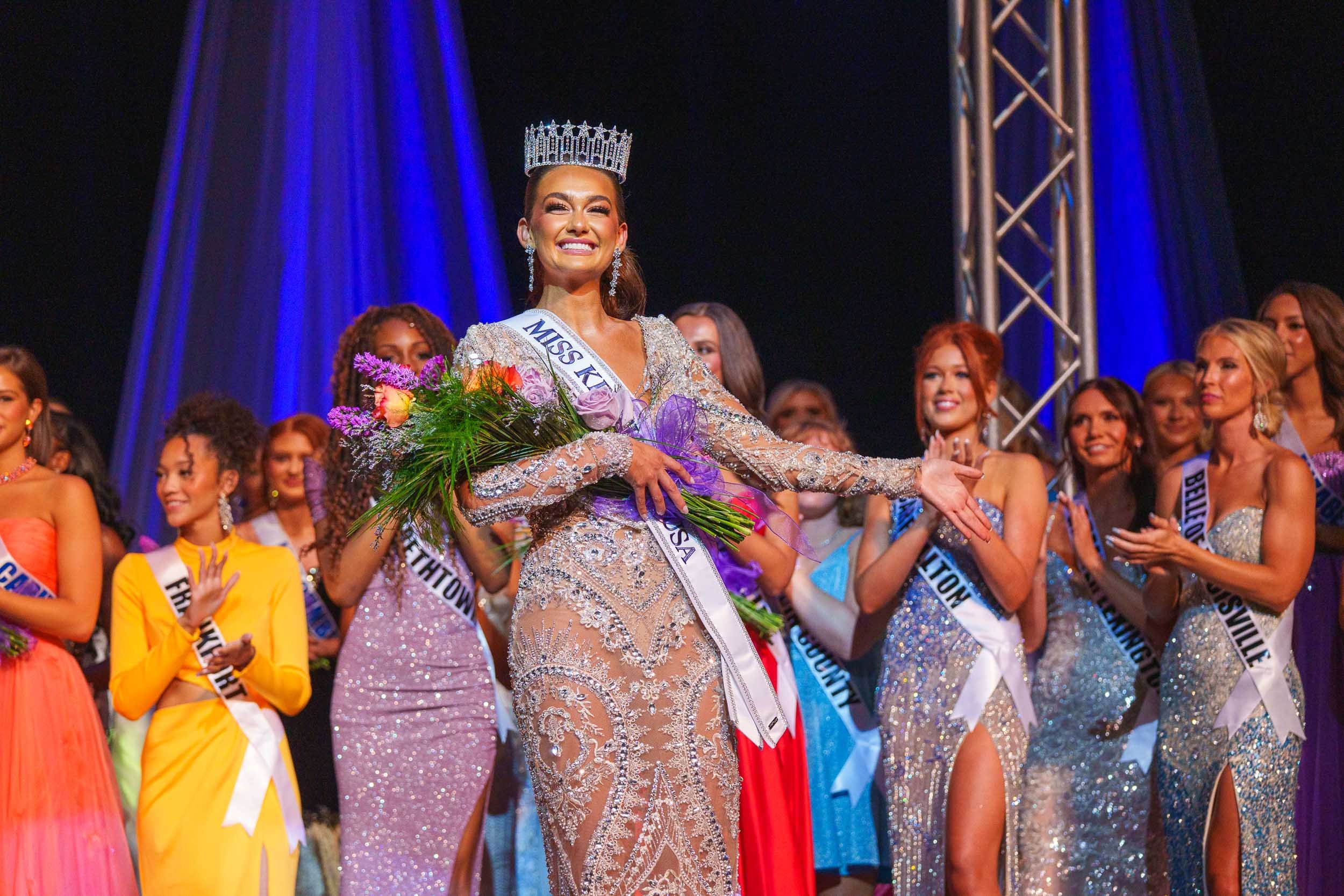 Miss Kentucky 2025 crowned at a beauty pageant, standing with a bouquet of flowers, wearing a tiara and a sash, surrounded by other contestants in evening gowns.