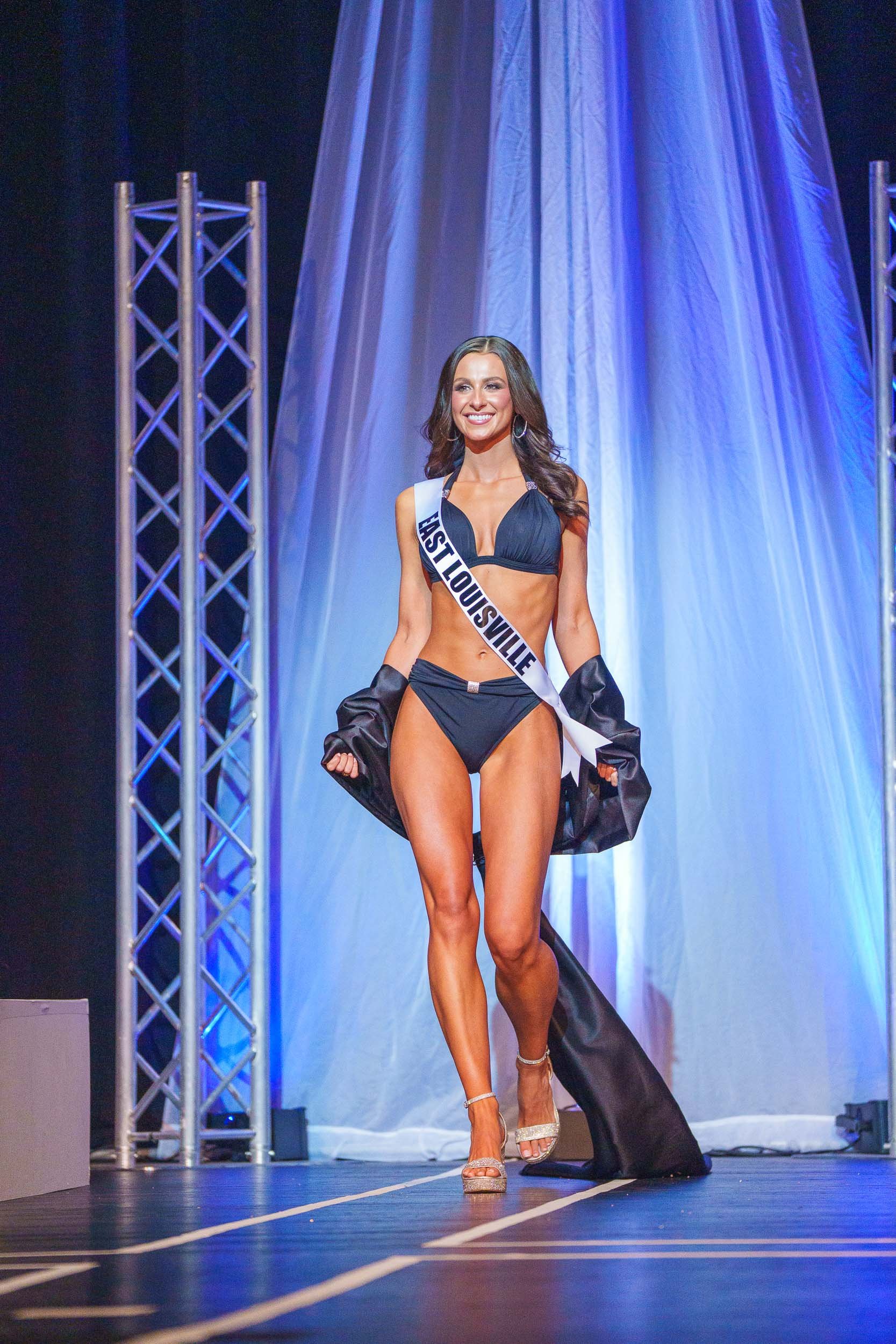 Miss USA contestant walking on stage during a beauty pageant, wearing a sash labeled 'Louisville,' black bikini, high heels, and holding a black dress. She is smiling, with stage lighting and a backdrop behind her.