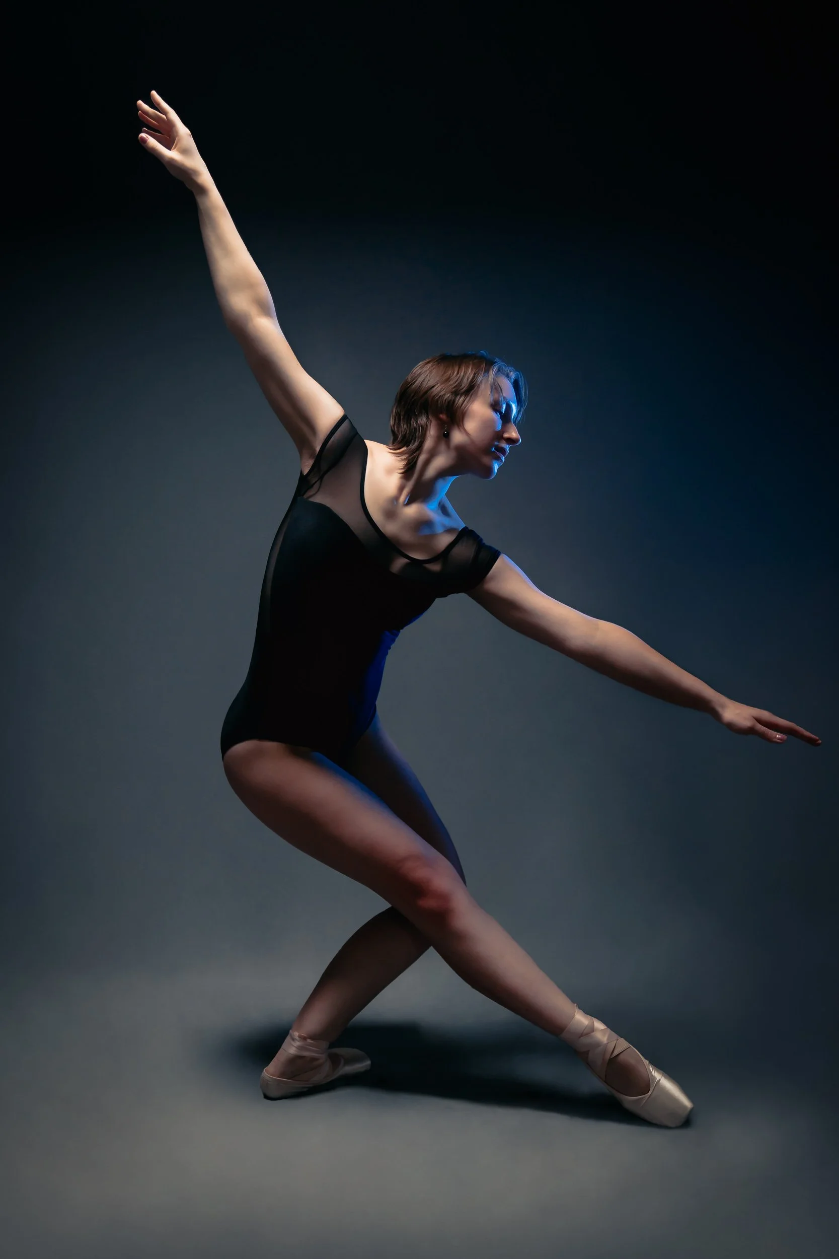 Ballet dancer with short brown hair practicing in a black leotard, posing on pointe with arms extended, against a dark gradient background.