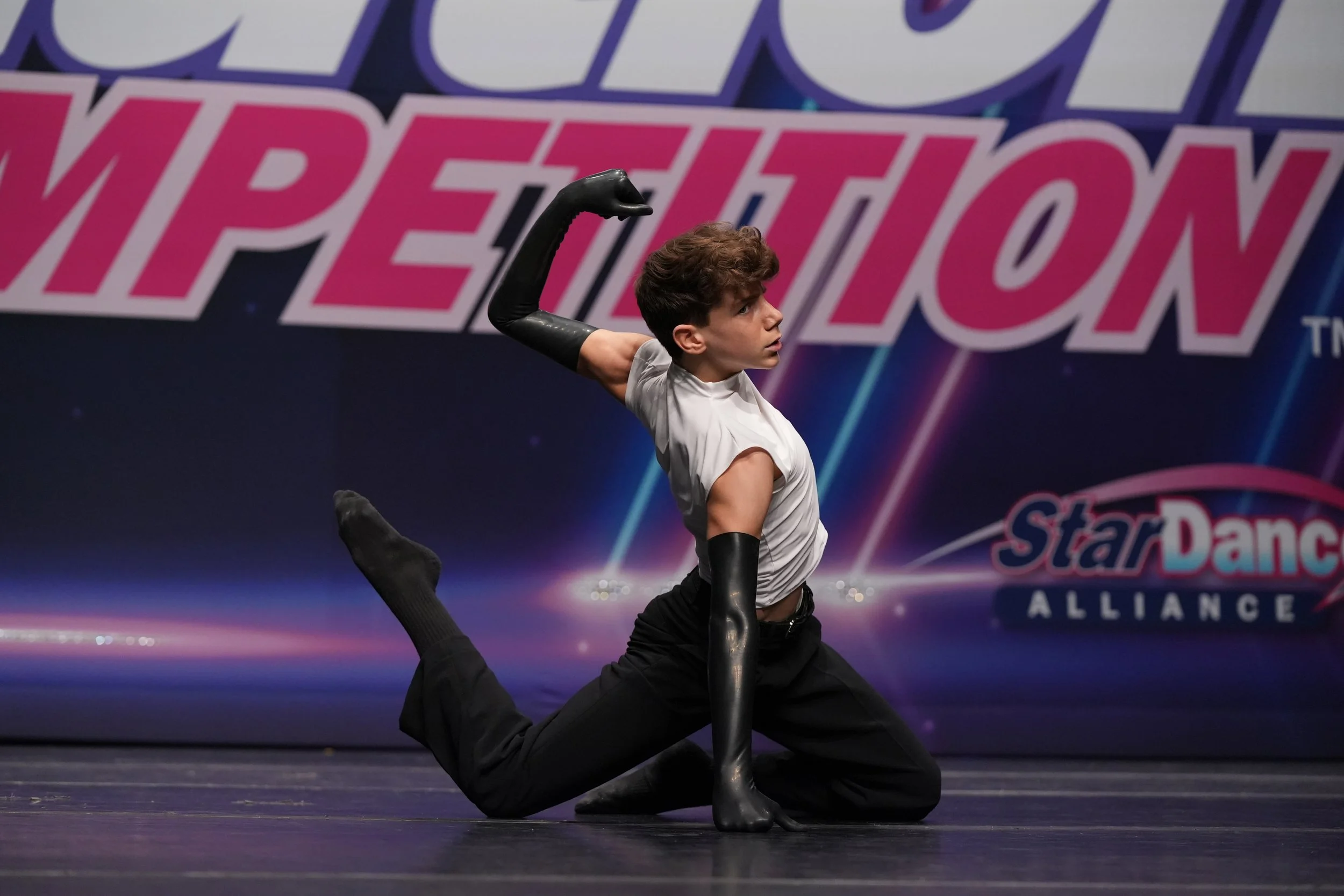 A male dancer performing on stage at Star Dance Alliance competition, in a pose with one knee on the floor, one arm bent with the hand near the head, and the other arm extended outward, wearing a white sleeveless top, black gloves, and black pants.