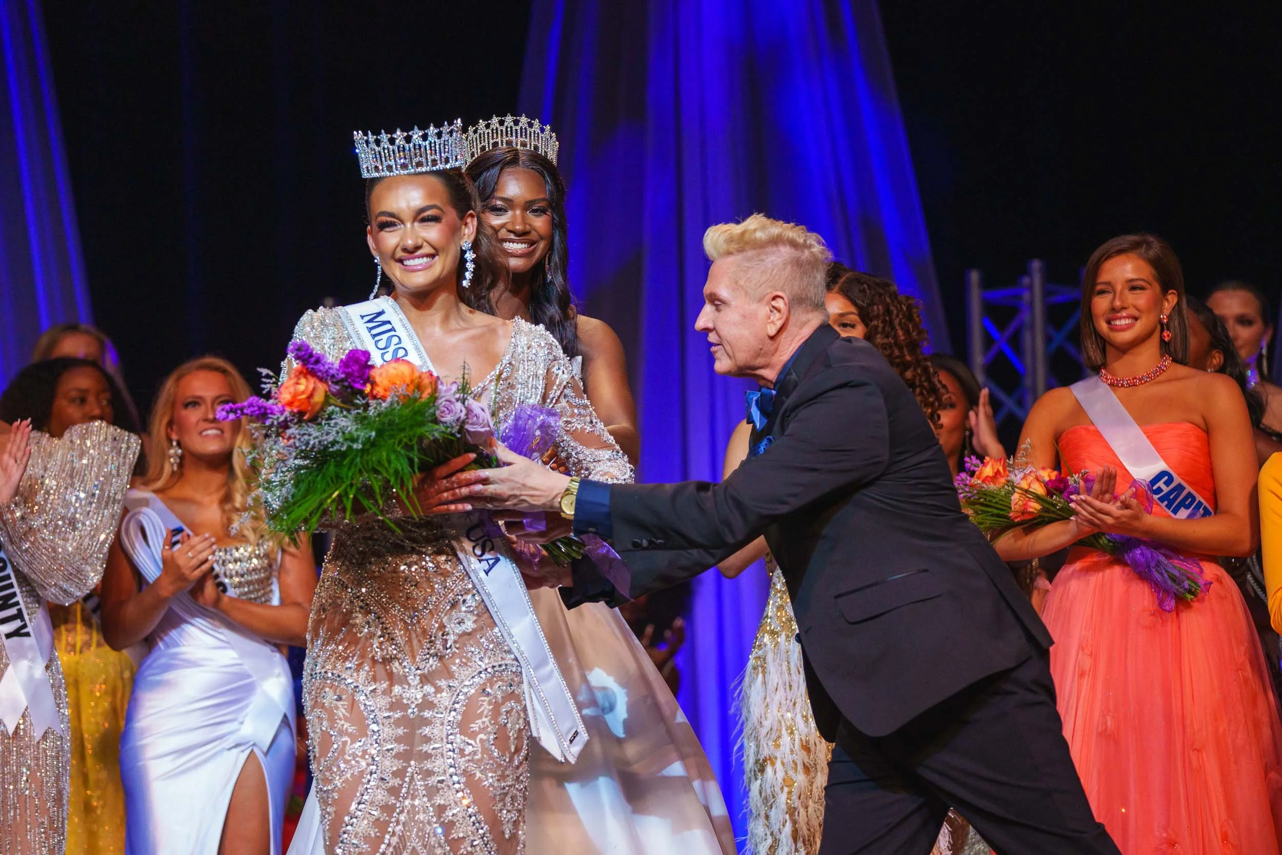 Miss Kentucky 2025 wearing a crown and sash, holding flowers, receiving an award on stage surrounded by other winners in evening gowns.