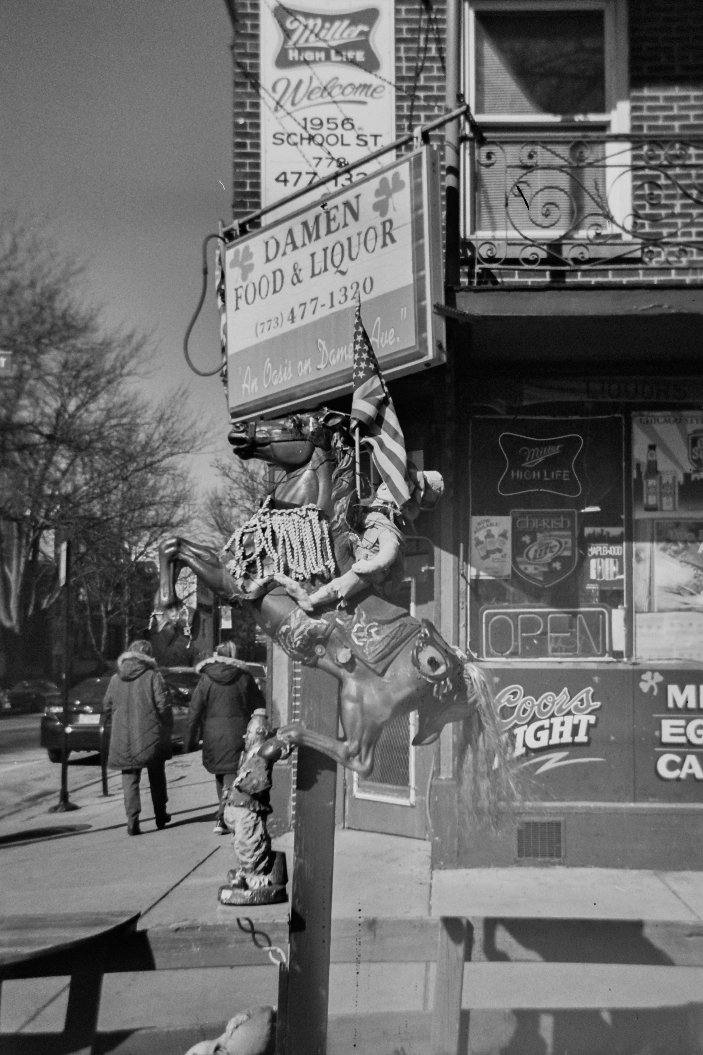 A man on a decorated horse statue on a city sidewalk, with a storefront and signs advertising food and liquor items in the background. Several pedestrians are visible walking past.