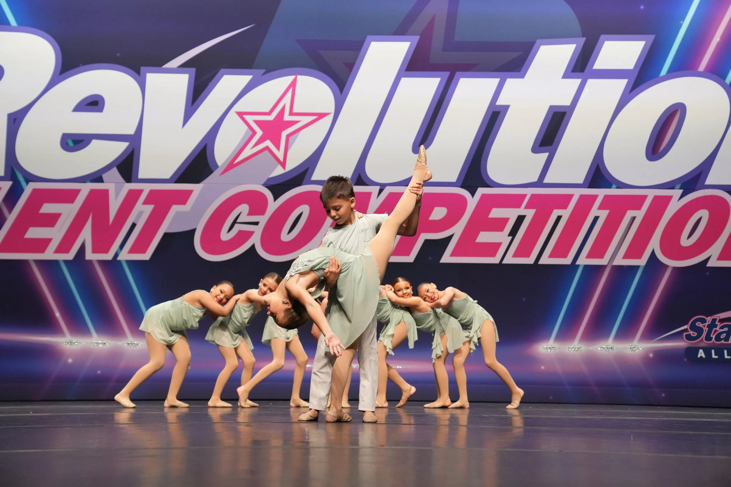 A ballet dance performance with seven children on stage, five girls in matching outfits and a boy lifting one girl. The background has a large, colorful sign reading 'Revolution Talent Competition'.