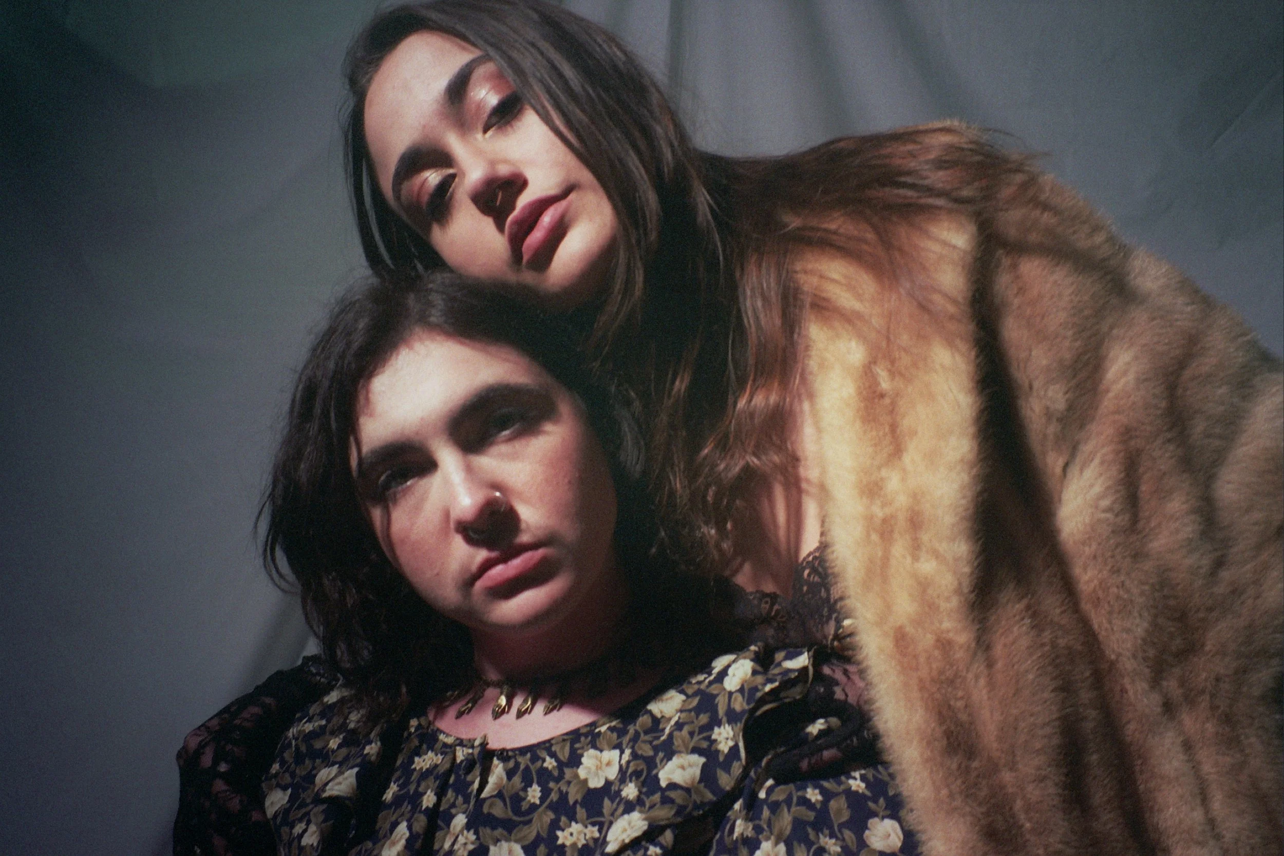 Close-up of two women with dark hair, one with a fur coat, posing closely together against a dark background.