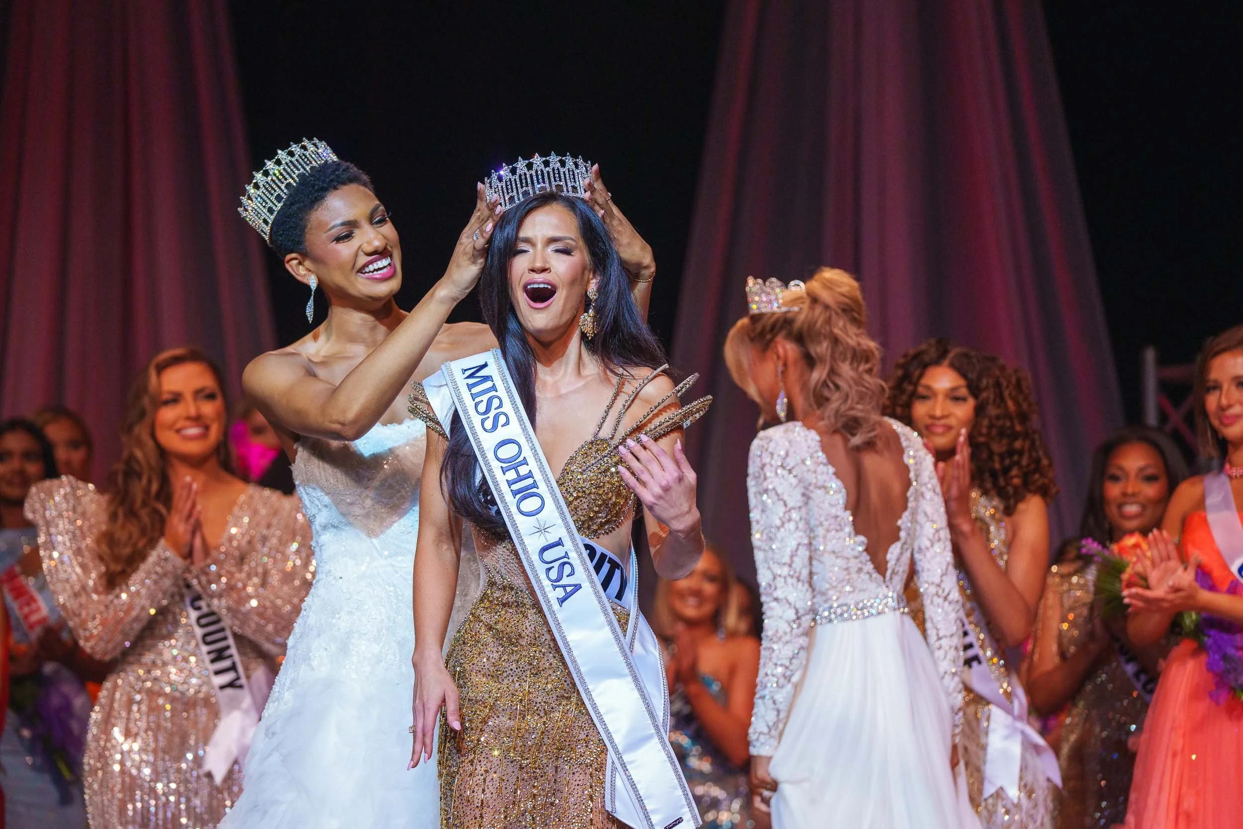 Miss Ohio 2025 USA crowned at a beauty pageant, surrounded by contestants in evening gowns, on stage with curtains in background.