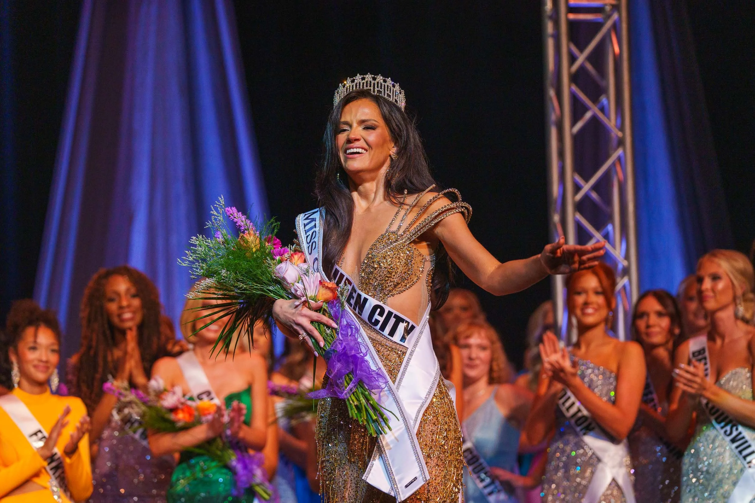 Miss Ohio 2025 wearing a crown and a sash that reads 'Miss City' is smiling and holding a bouquet of flowers during a beauty pageant. She is dressed in a sparkling gown and is on stage with other contestants visible in the background.