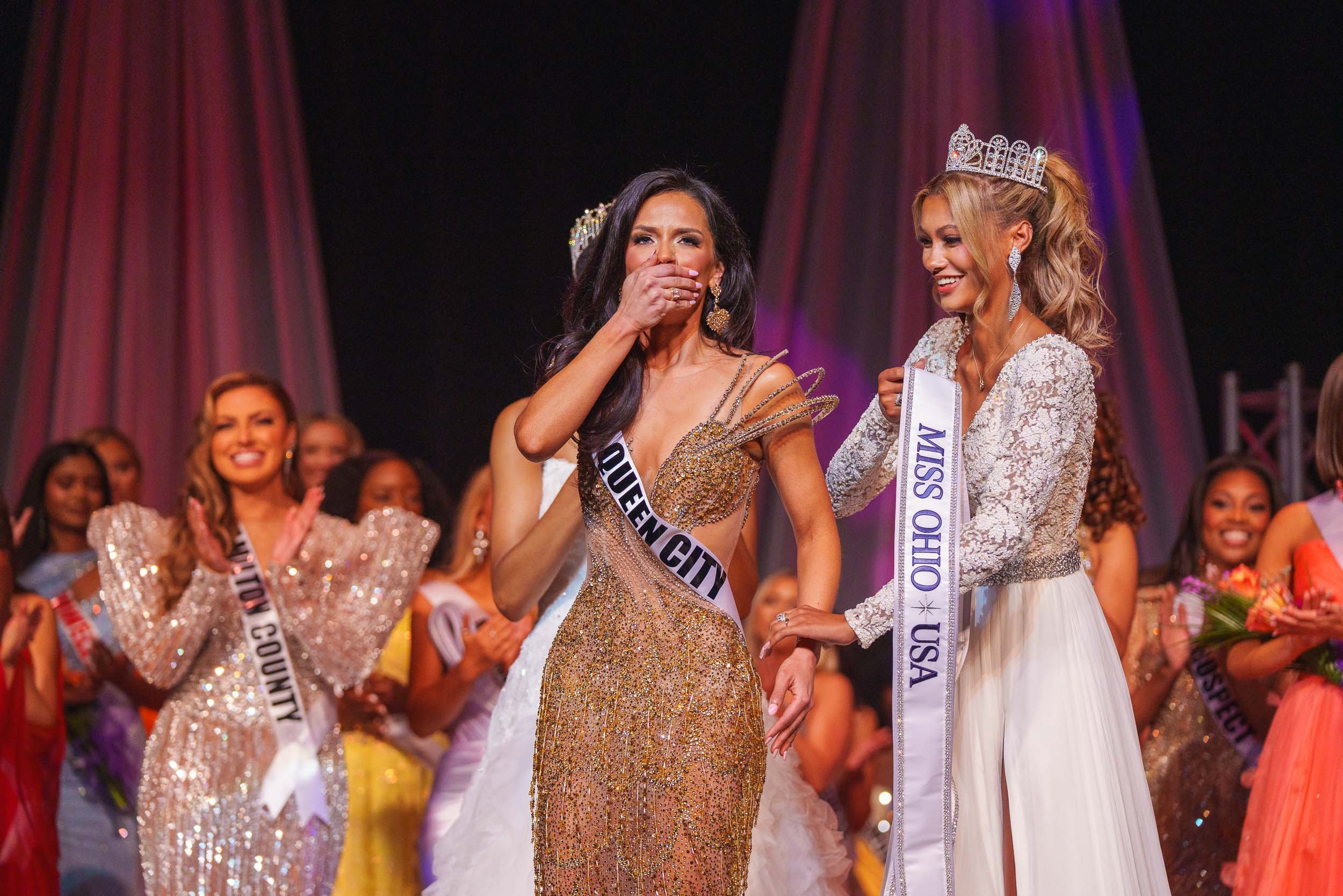 Two women wearing crowns and sashes at a beauty pageant, waving and smiling, with several other women clapping and smiling in the background.