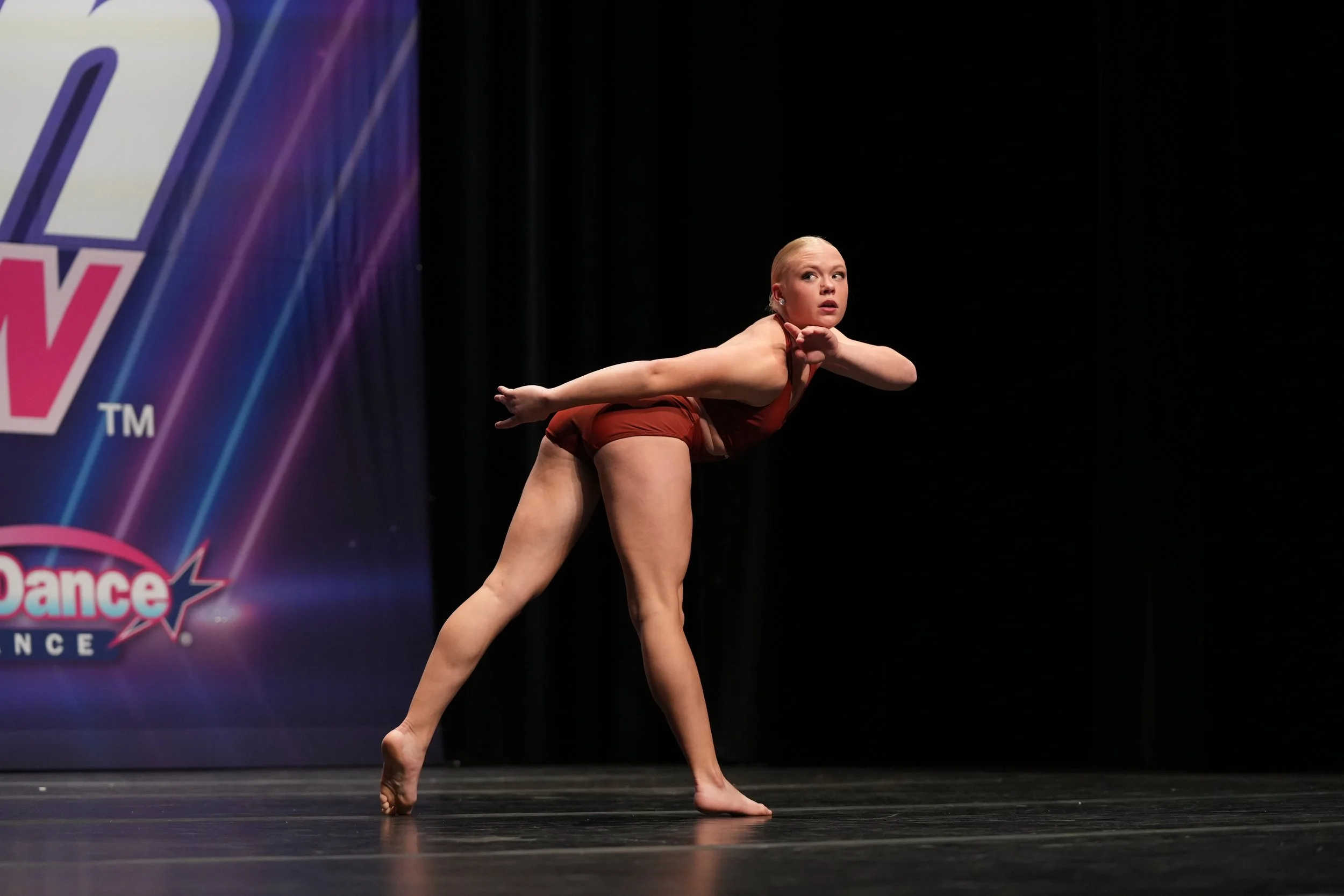 A young woman with blond hair, wearing a maroon dance costume, performing a dance pose on stage during a dance competition, with a dark background and a colorful stage banner to the left.