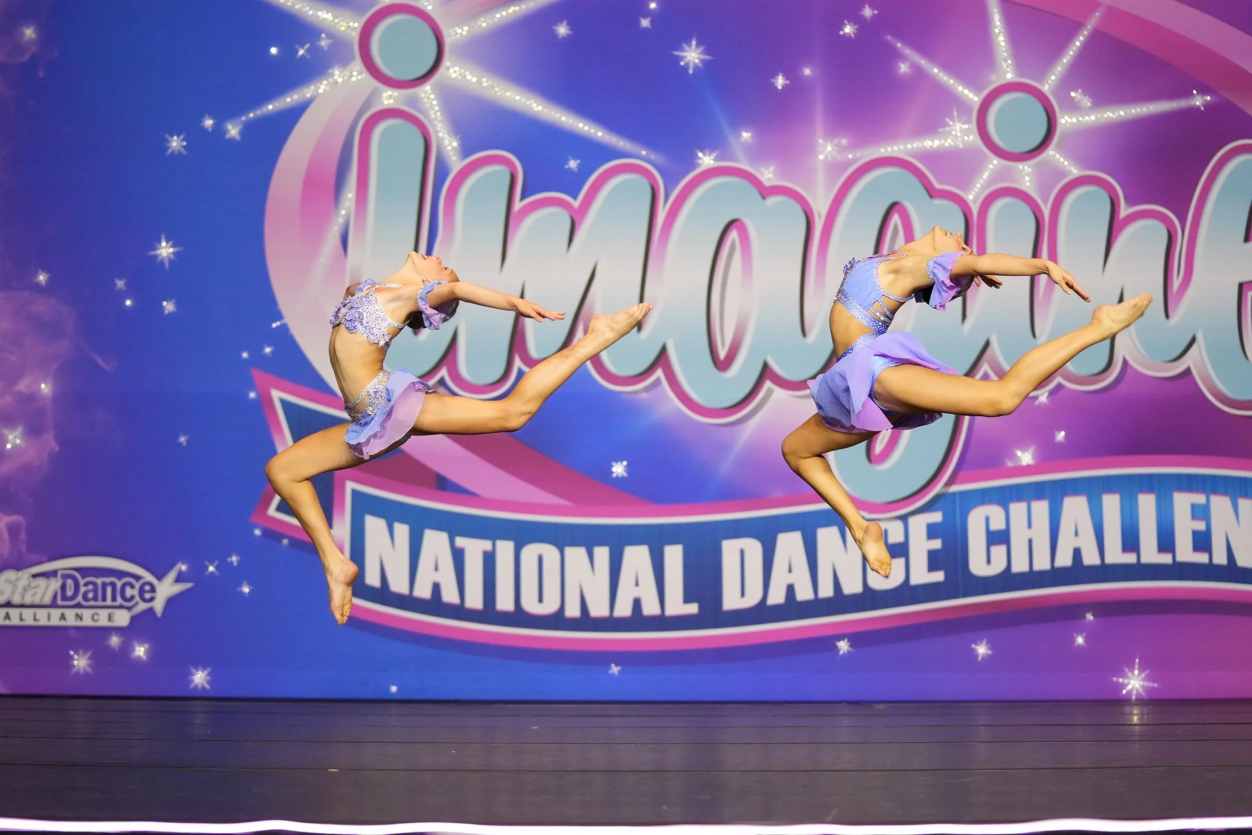 Two young female dancers in purple costumes performing a leap in mid-air on stage at a national dance competition, with a colorful backdrop reading 'Star Dance Alliance National Dance Challenge'.