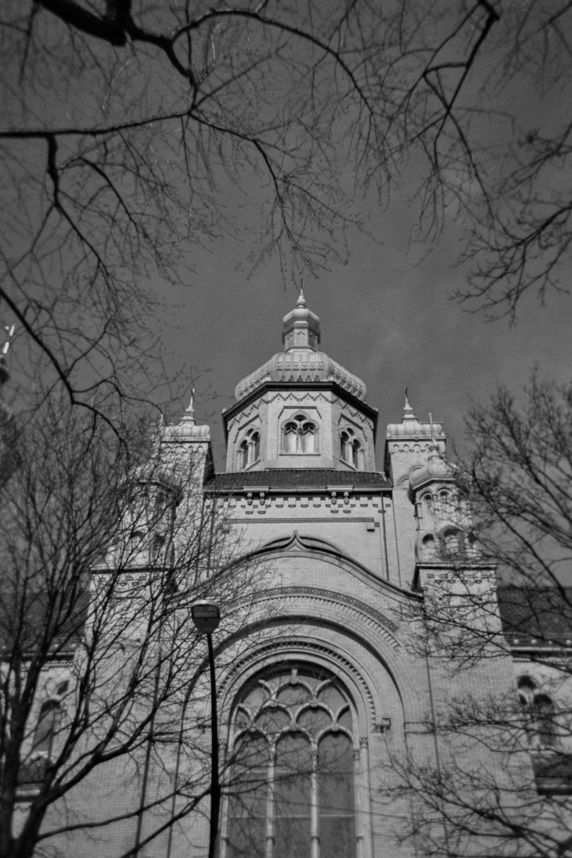 A black and white photo of a church with Gothic architectural details, trees without leaves in the foreground, and cloudy night sky.