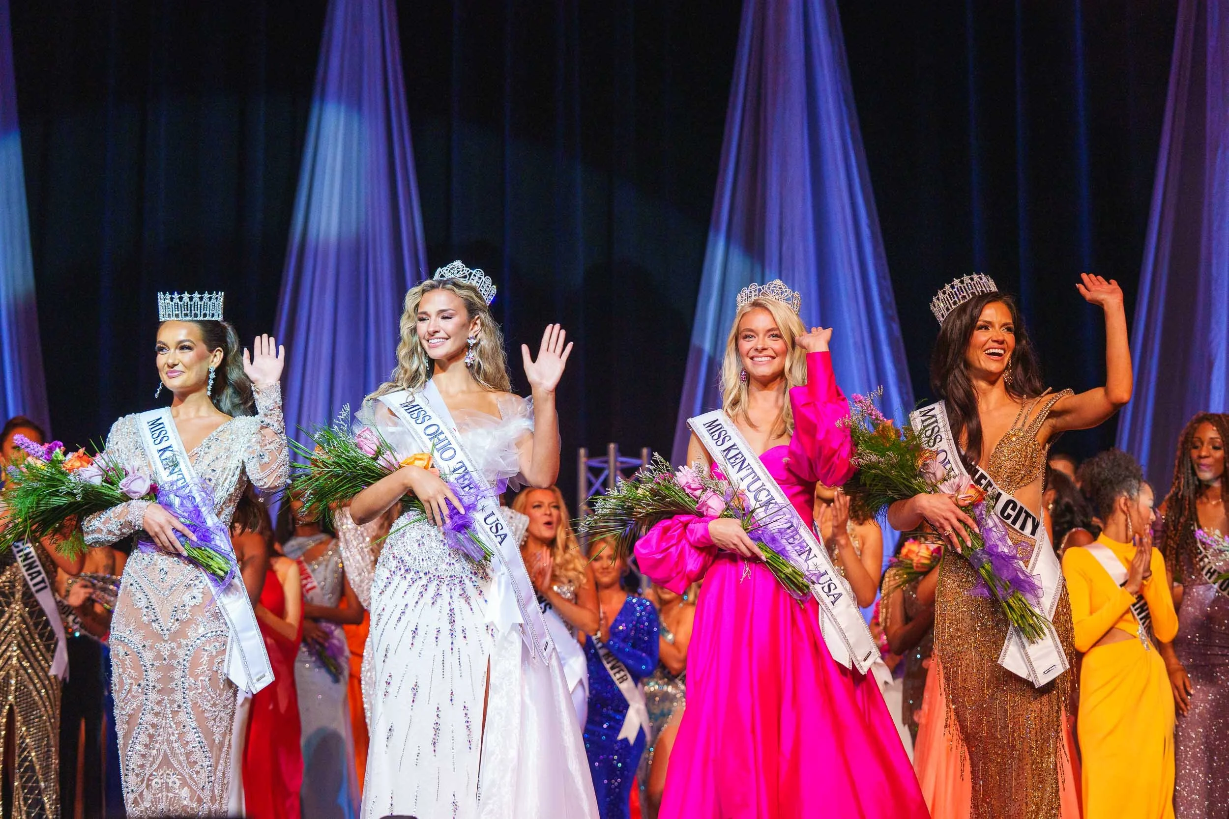 Four women on stage wearing crowns and sashes, holding bouquets, celebrating at a beauty pageant. The women are dressed in elegant evening gowns, and the background features draped curtains and stage lighting.
