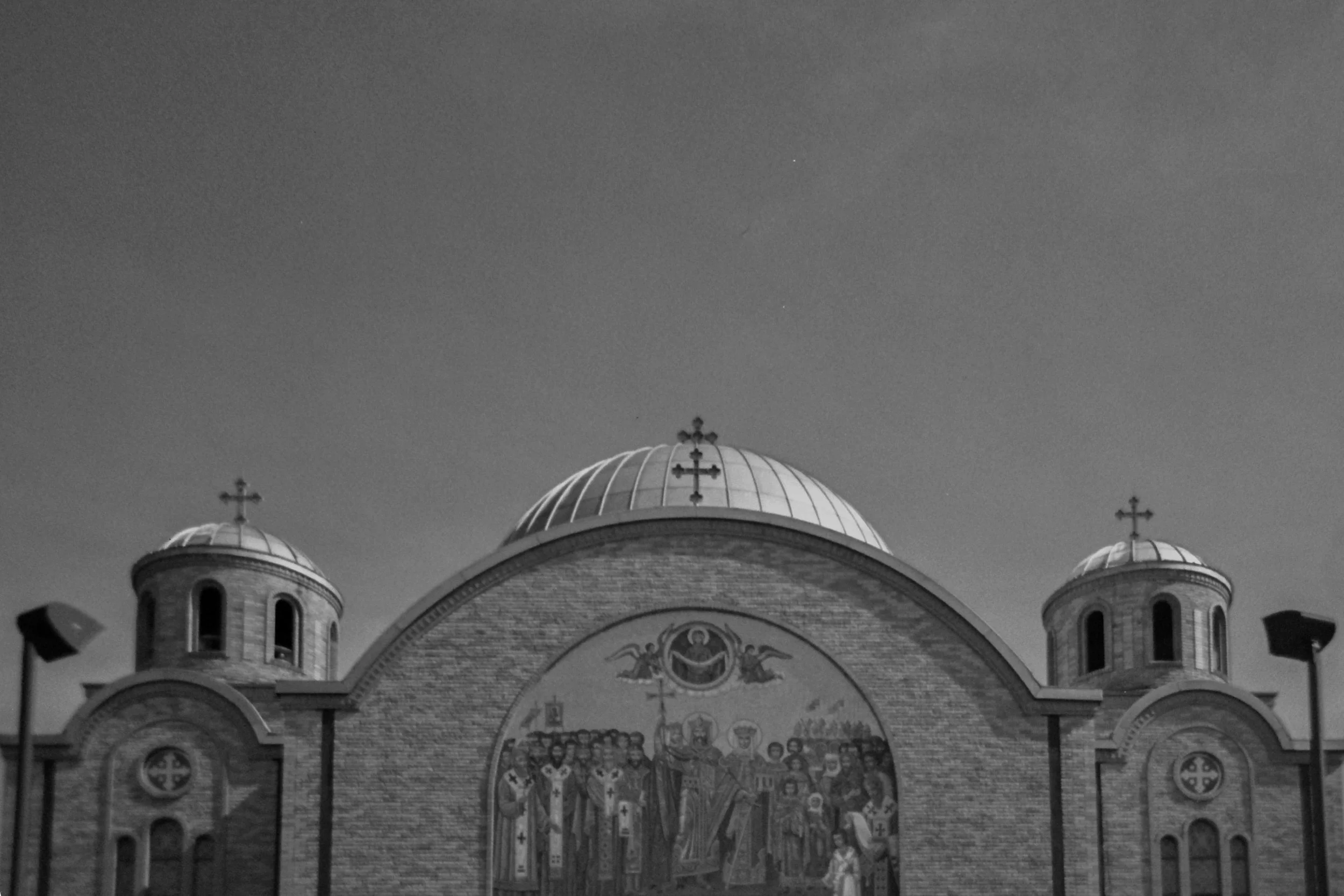 Black and white photograph of a church with three domes and crosses on top, featuring colorful religious icons and murals on the front facade.