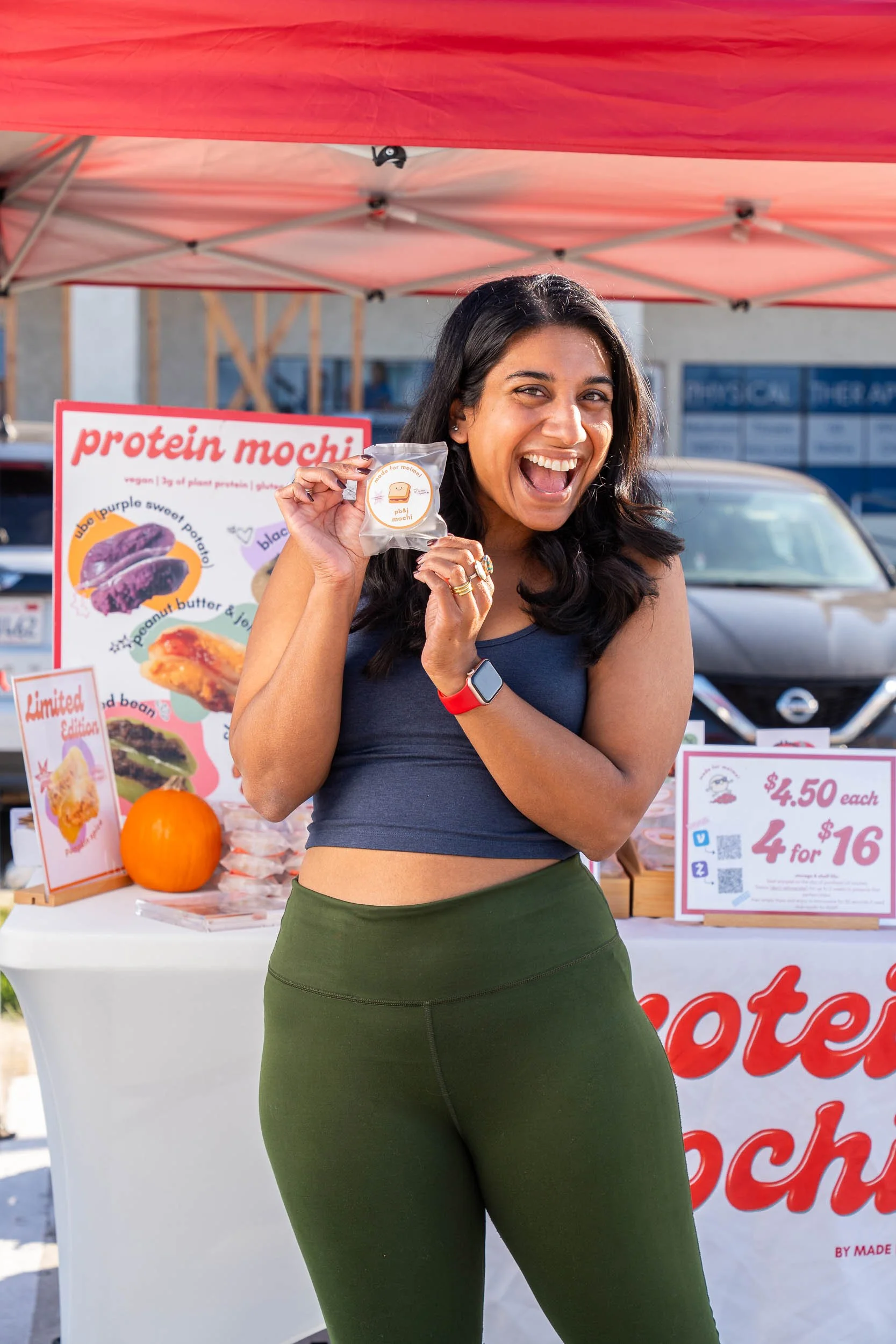 A woman happily holding a packaged protein mochi at a booth with a sign displaying the product and its prices, under a pink canopy outdoors.