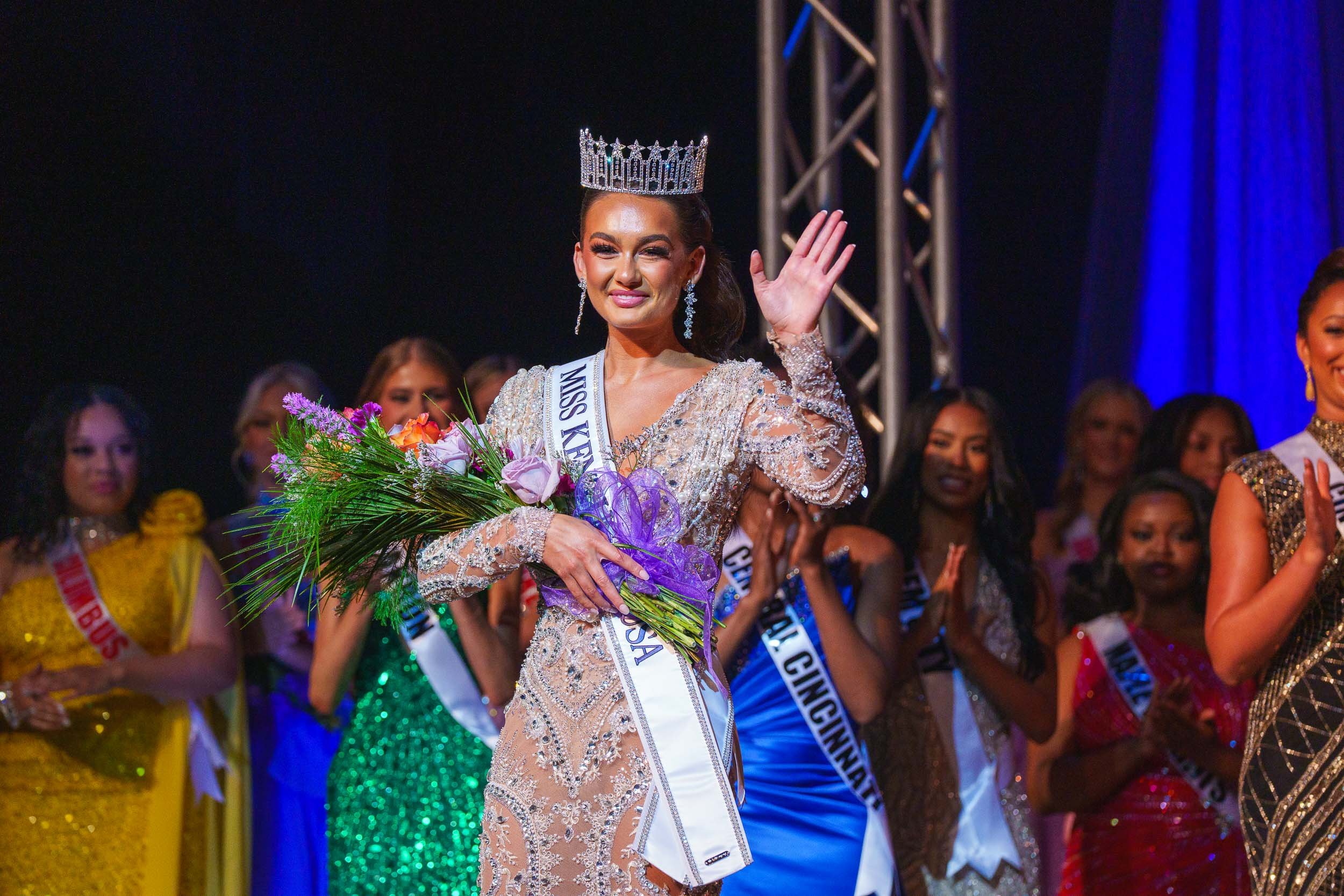 Miss Kentucky 2025 winner wearing a crown and sash, holding a bouquet, waving to the audience at a beauty pageant stage with other contestants in colorful dresses behind her.