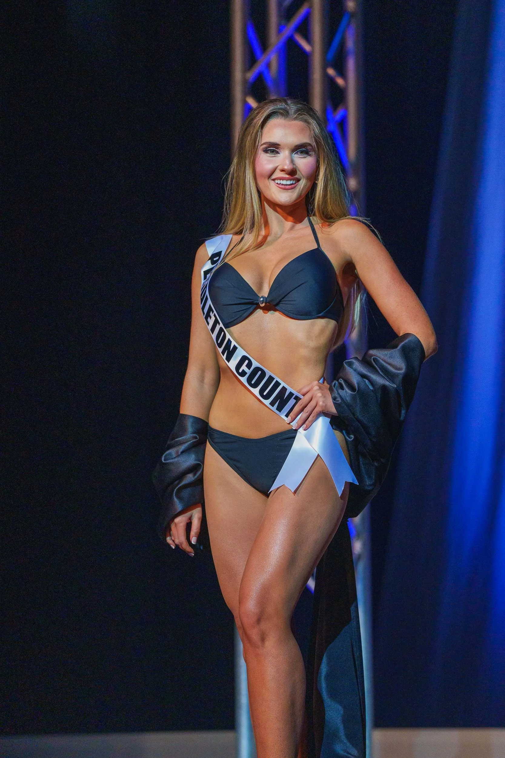A woman wearing a black bikini and a sash that says 'PENNSYLVANIA COUNTY' is on stage during a beauty pageant. She has long, styled blonde hair, bright makeup, and a confident smile, with stage lighting and a metallic truss in the background.