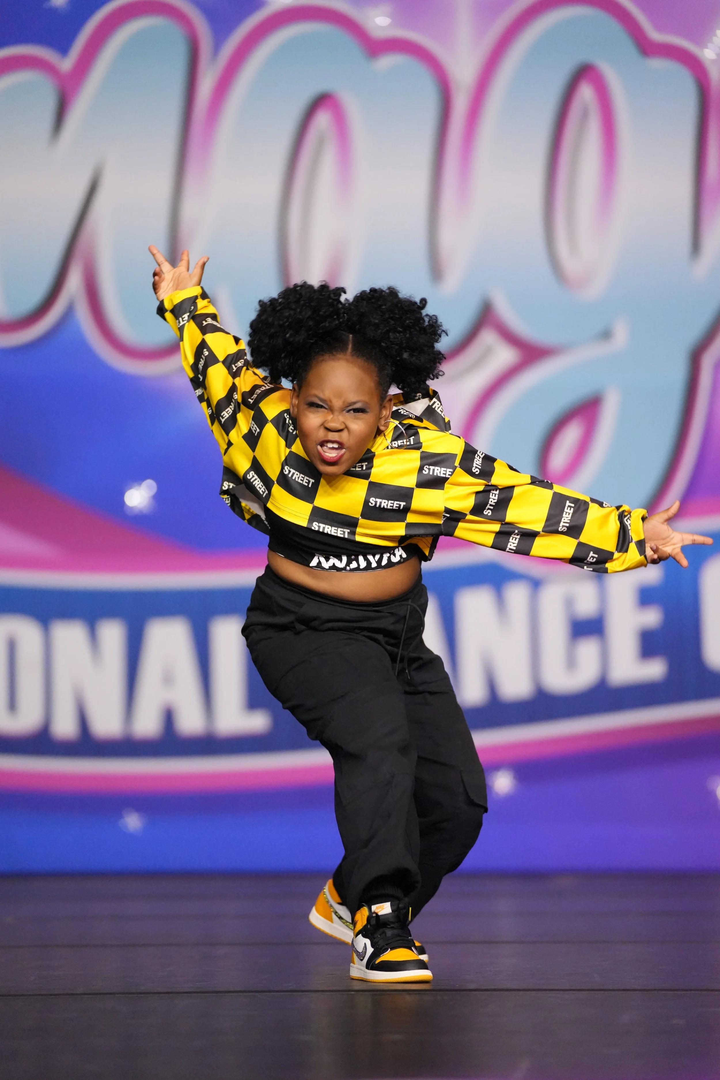 A young girl with curly hair in a yellow and black checkered jacket, black pants, and sneakers, dancing on stage during a dance competition.